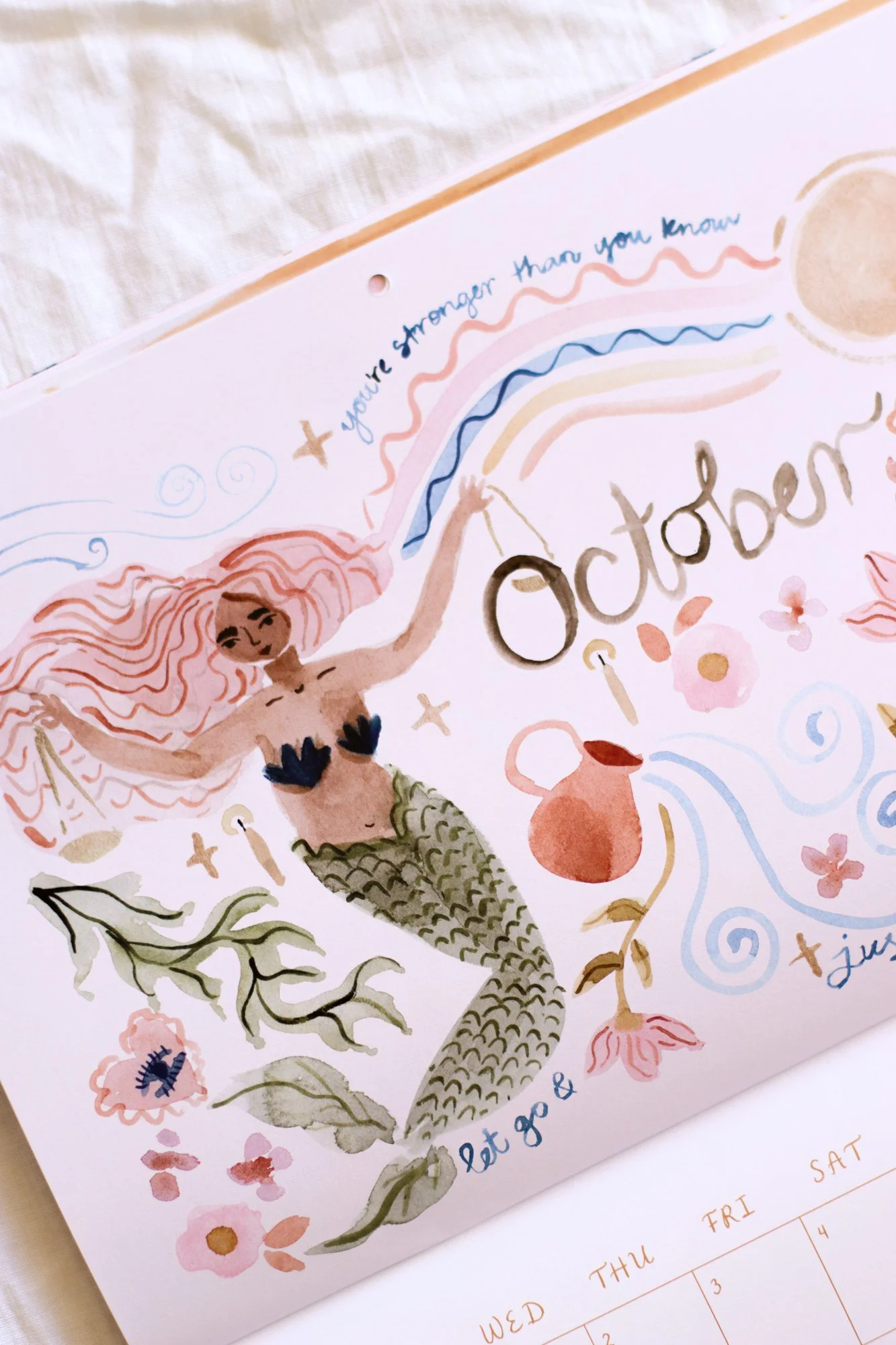 A colorful illustrated calendar page featuring a mermaid holding a rainbow and surrounded by flowers, leaves, and sea creatures, with handwritten notes and dates.