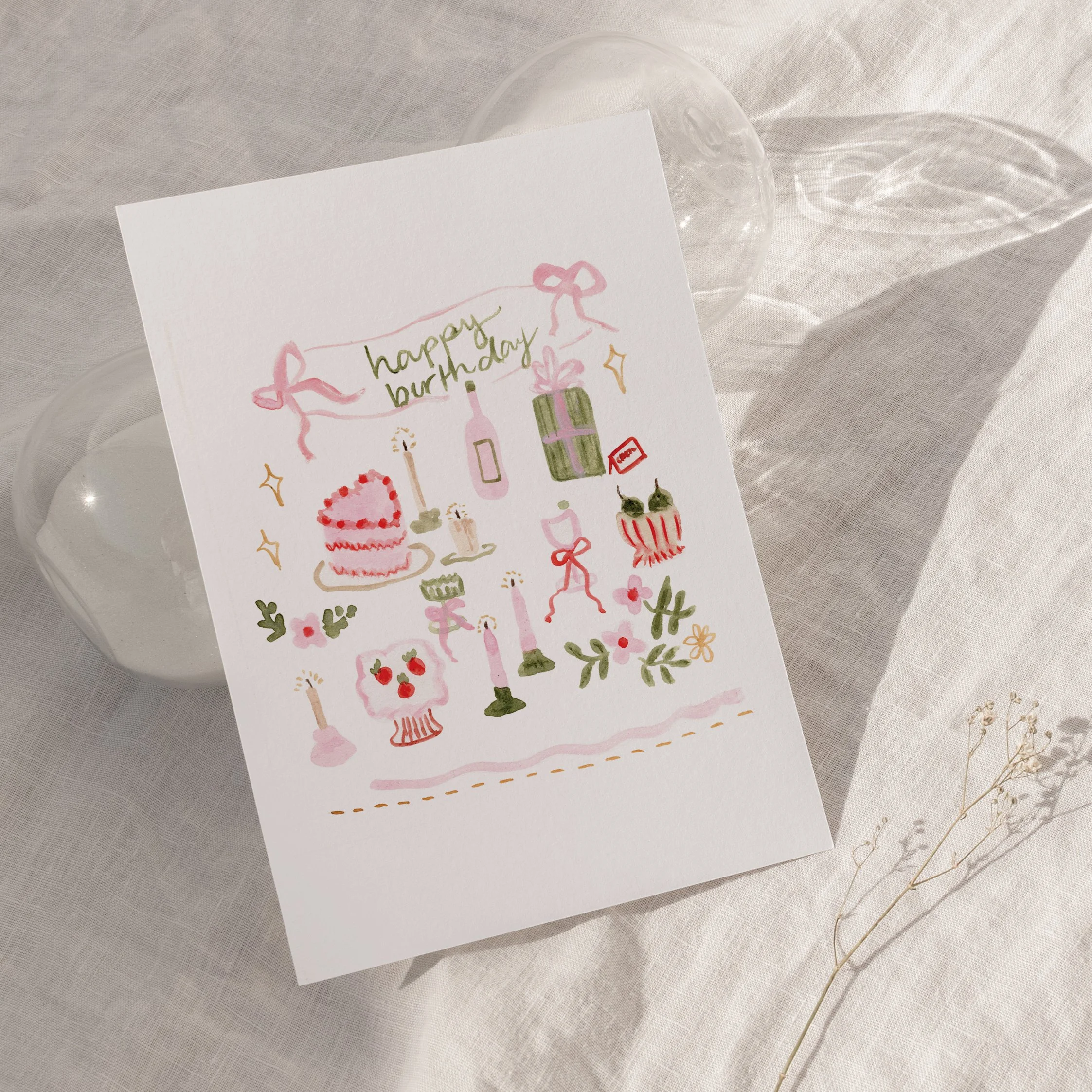 A white greeting card with pink, green, orange, and red decorative drawings and the words 'happy birthday' written in green, partially covered by a transparent glass bowl on a light fabric surface.