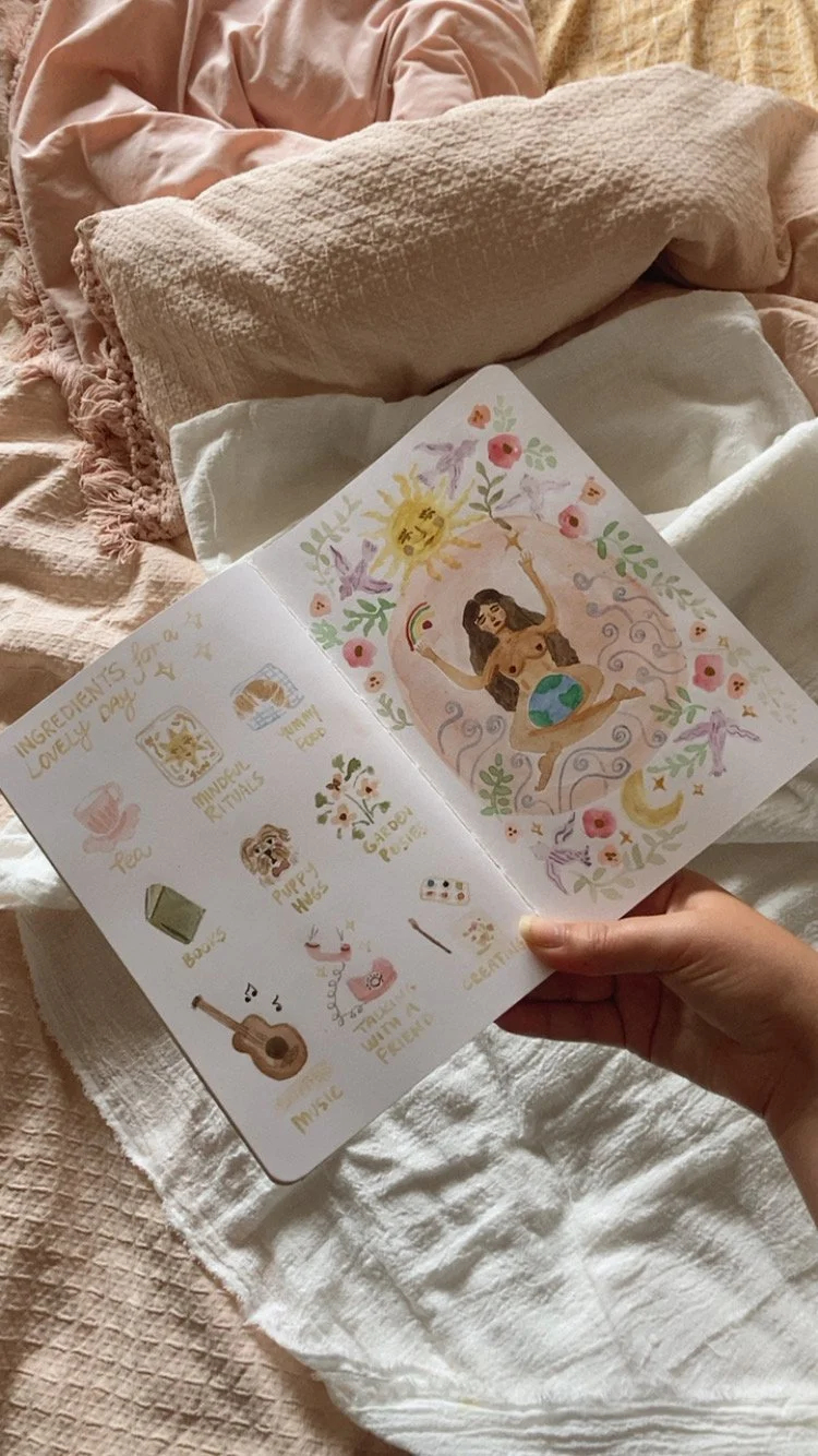 A hand holding an illustrated booklet open to a page with a drawing of a goddess figure surrounded by flowers and symbols, and a list of favorite ingredients for a love divination ritual, including tea, amber ritual, tarot, puppy paws, plant dew, a s