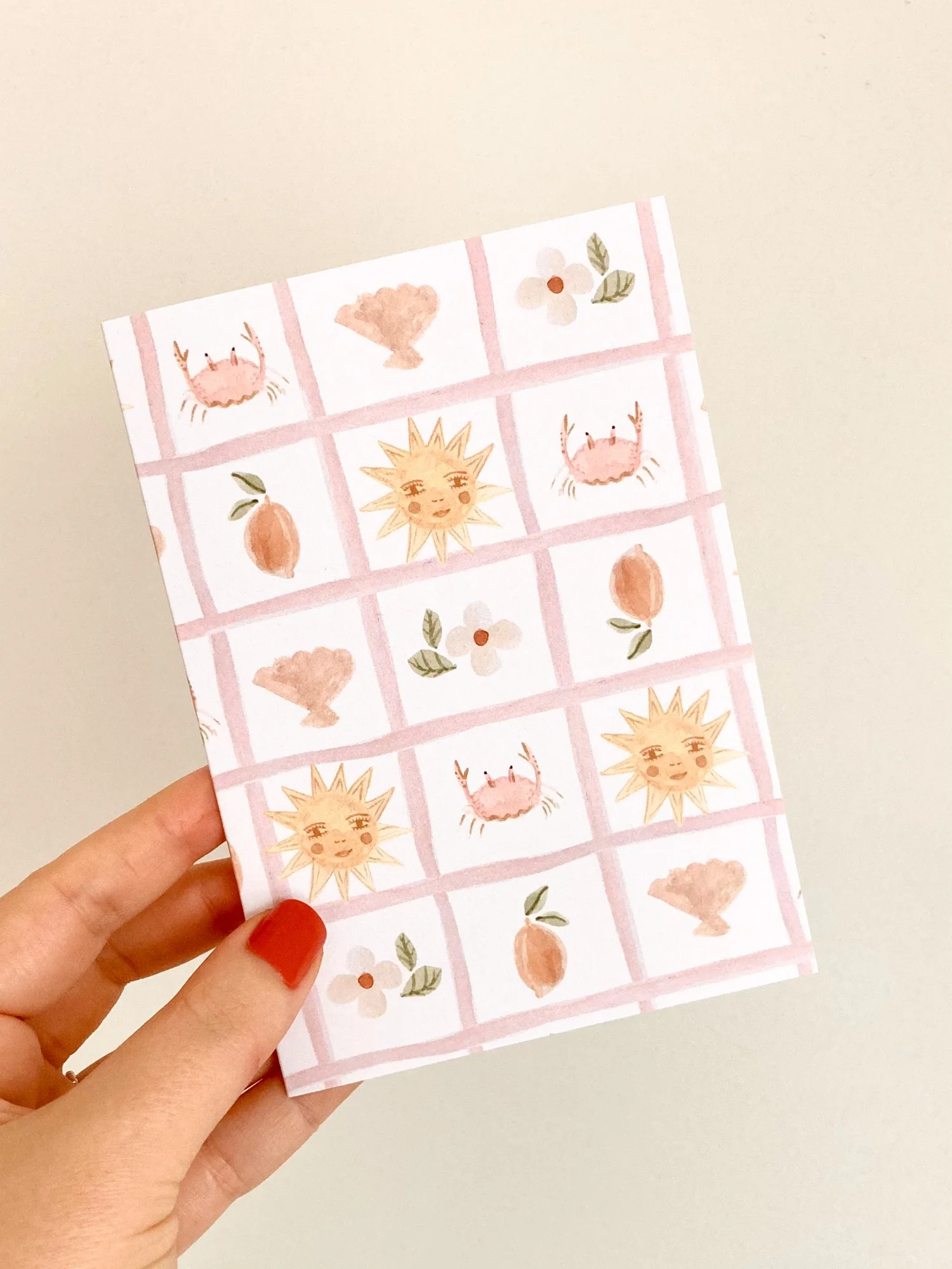 Person holding a sheet of paper with watercolor illustrations of crabs, flowers, a lemon, and a smiling sun, with a light-colored background.