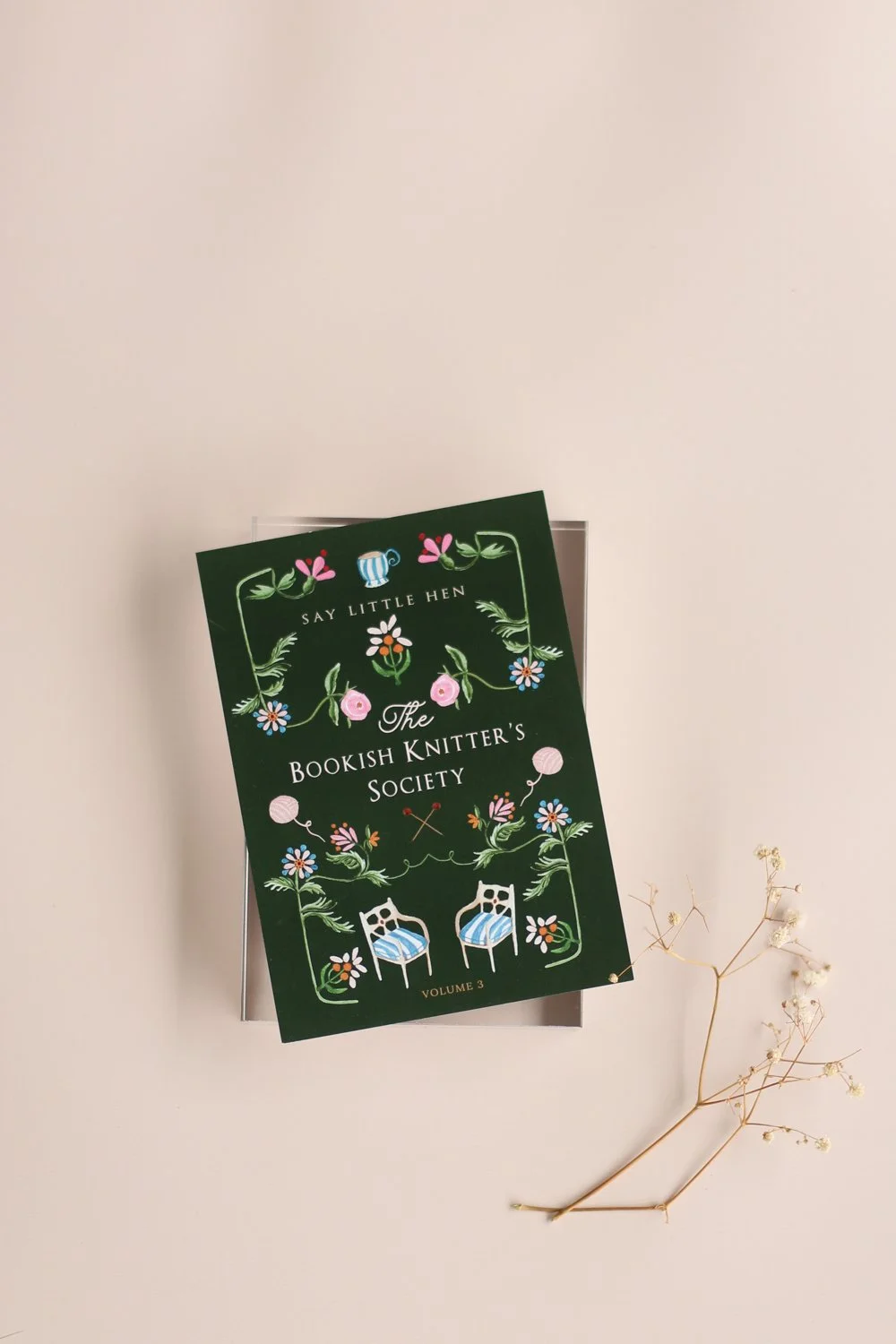 A book titled "The Bookish Knitter's Society" Volume 3, resting on top of a white envelope. The book cover is dark green with floral and knitting-themed illustrations, and there is a sprig of dried flowers placed beside it on a light-colored surface.