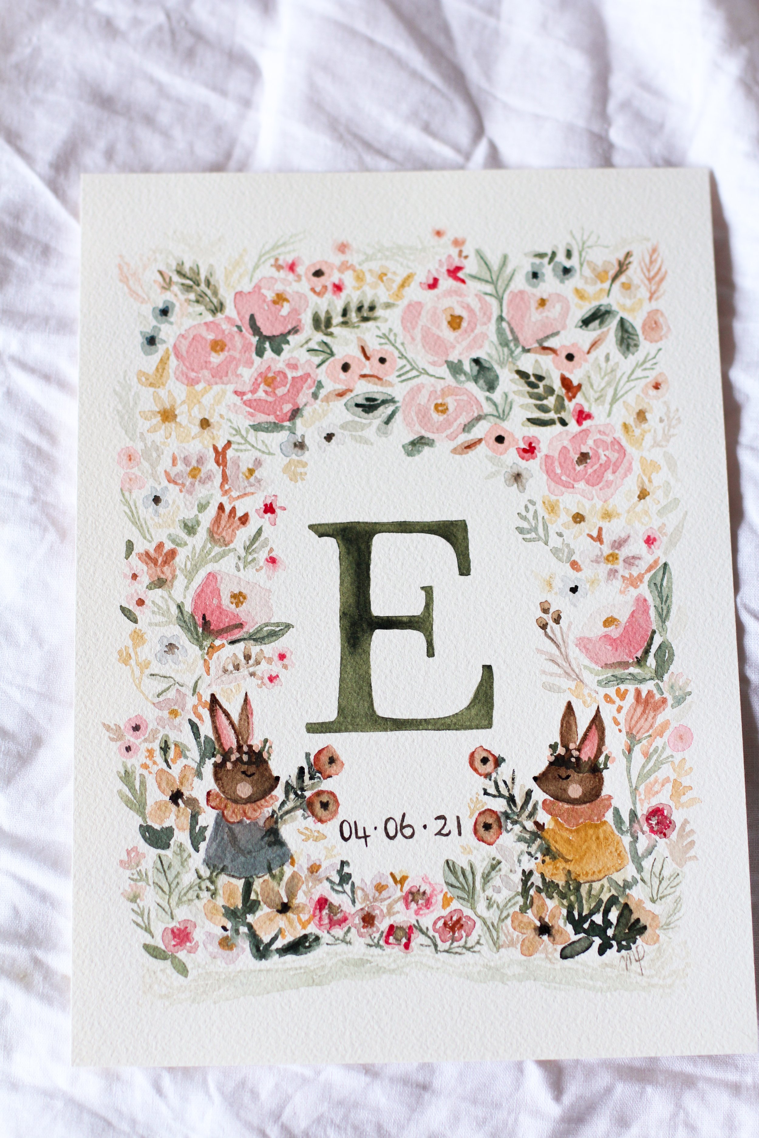 A decorative paper with a watercolor floral wreath surrounding a large letter 'E' and two illustrated rabbits at the bottom, with the date "04-06-21" written below.