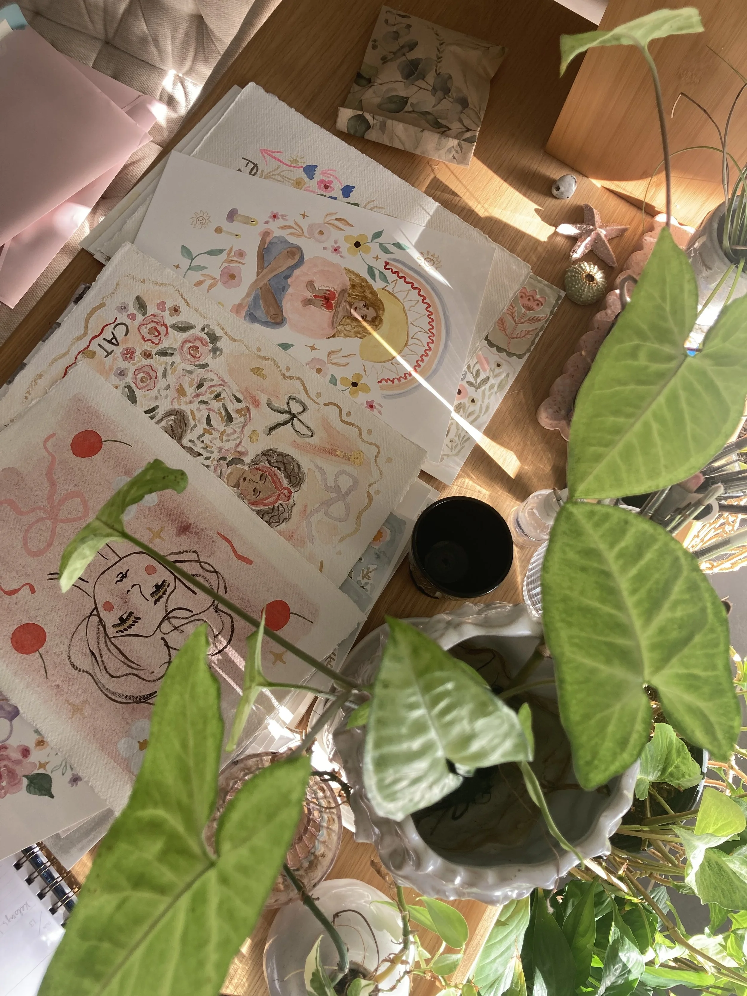 A wooden desk with potted plants, decorative notebooks, watercolor artwork, and stationery items, illuminated by natural sunlight.