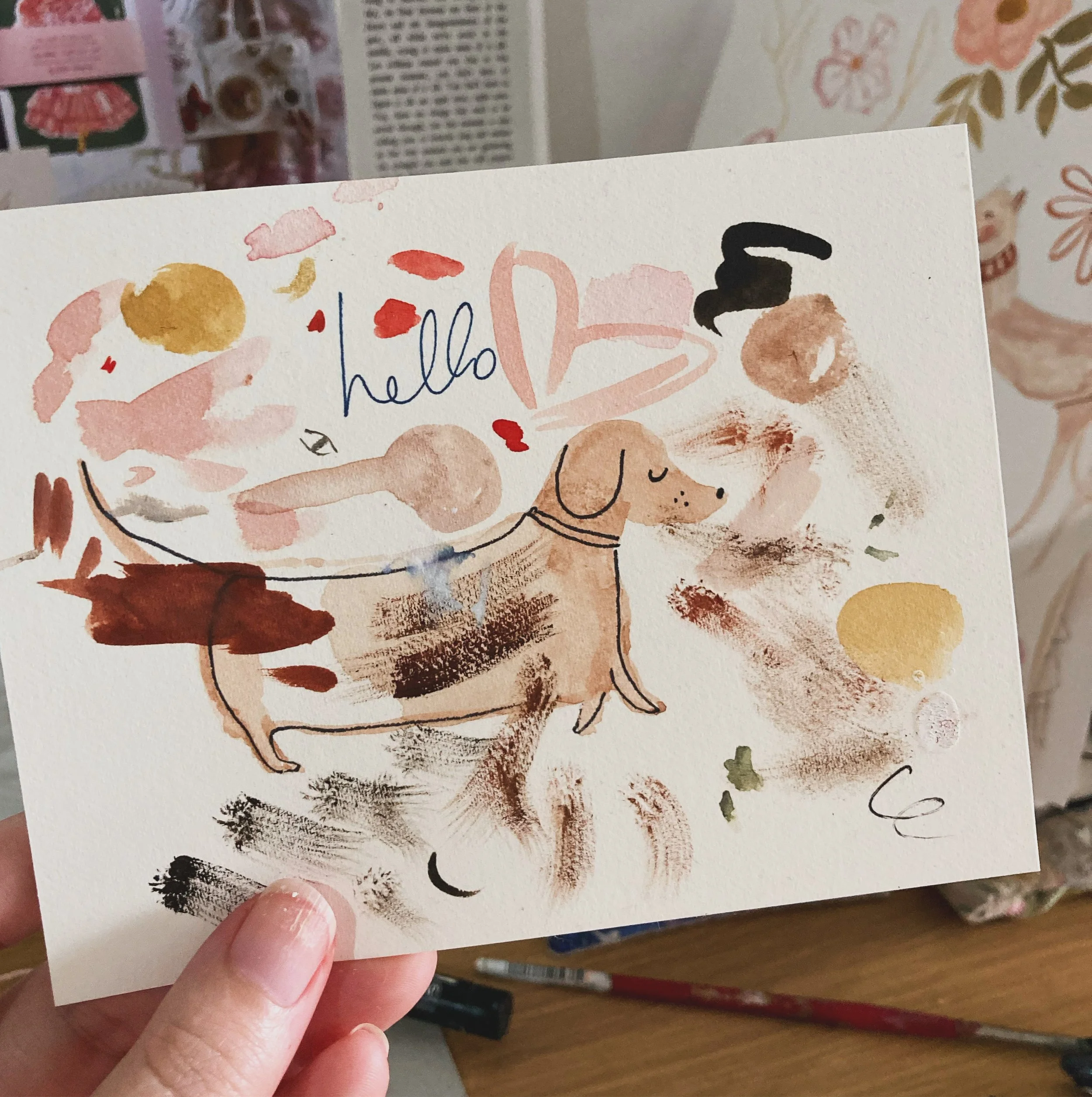 A person holding a watercolor painted greeting card with the word "hello" written on it, surrounded by abstract colorful shapes.