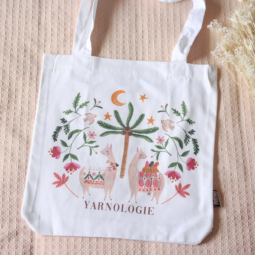 A white tote bag with a colorful illustration of two llamas carrying blankets, a palm tree, a crescent moon, stars, and various flowers, with the word "YARNOLOGIE" written at the bottom.