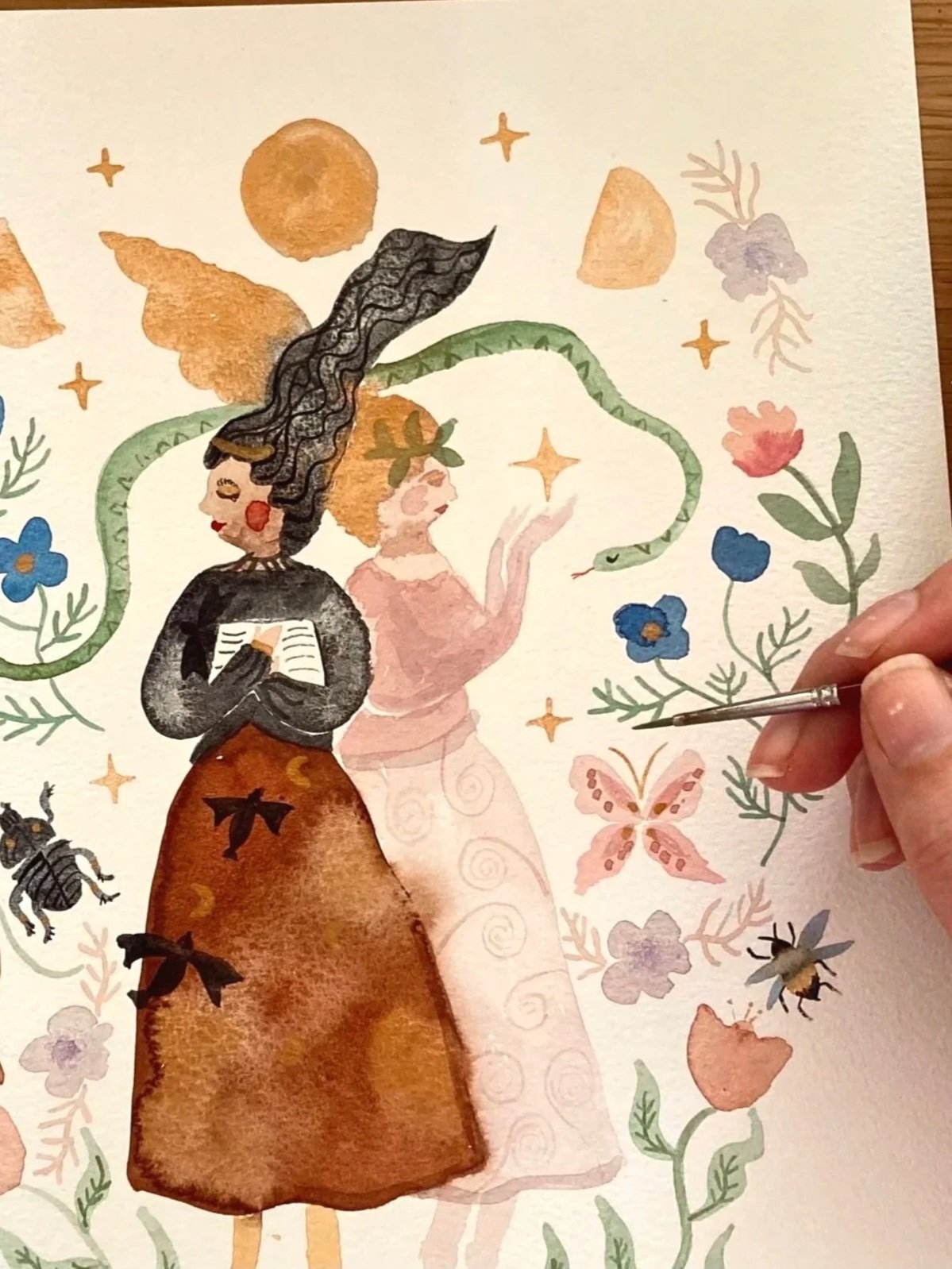 Watercolor illustration of two stylized women, surrounded by flowers, butterflies, bees, and stars, with a hand holding a brush adding details.