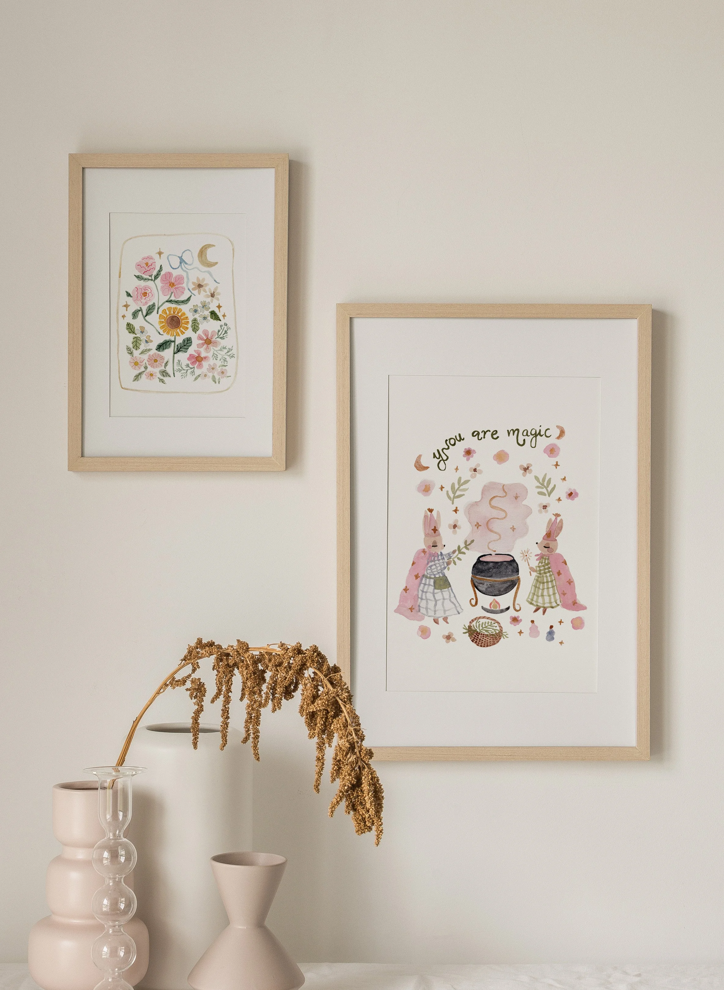 Two framed art prints hanging on a white wall; one features colorful flowers and celestial elements, the other depicts a whimsical scene with two bunnies around a cauldron and the phrase "you are magic." In front, a dried plant arrangement on a table with pastel-colored vases and a glass vessel.