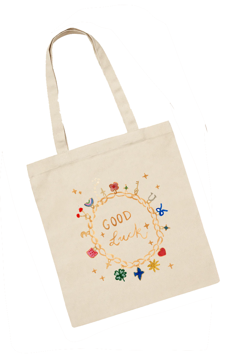 A cream-colored tote bag with colorful charms and the words 'Good Luck' written inside a chain circle on the front.