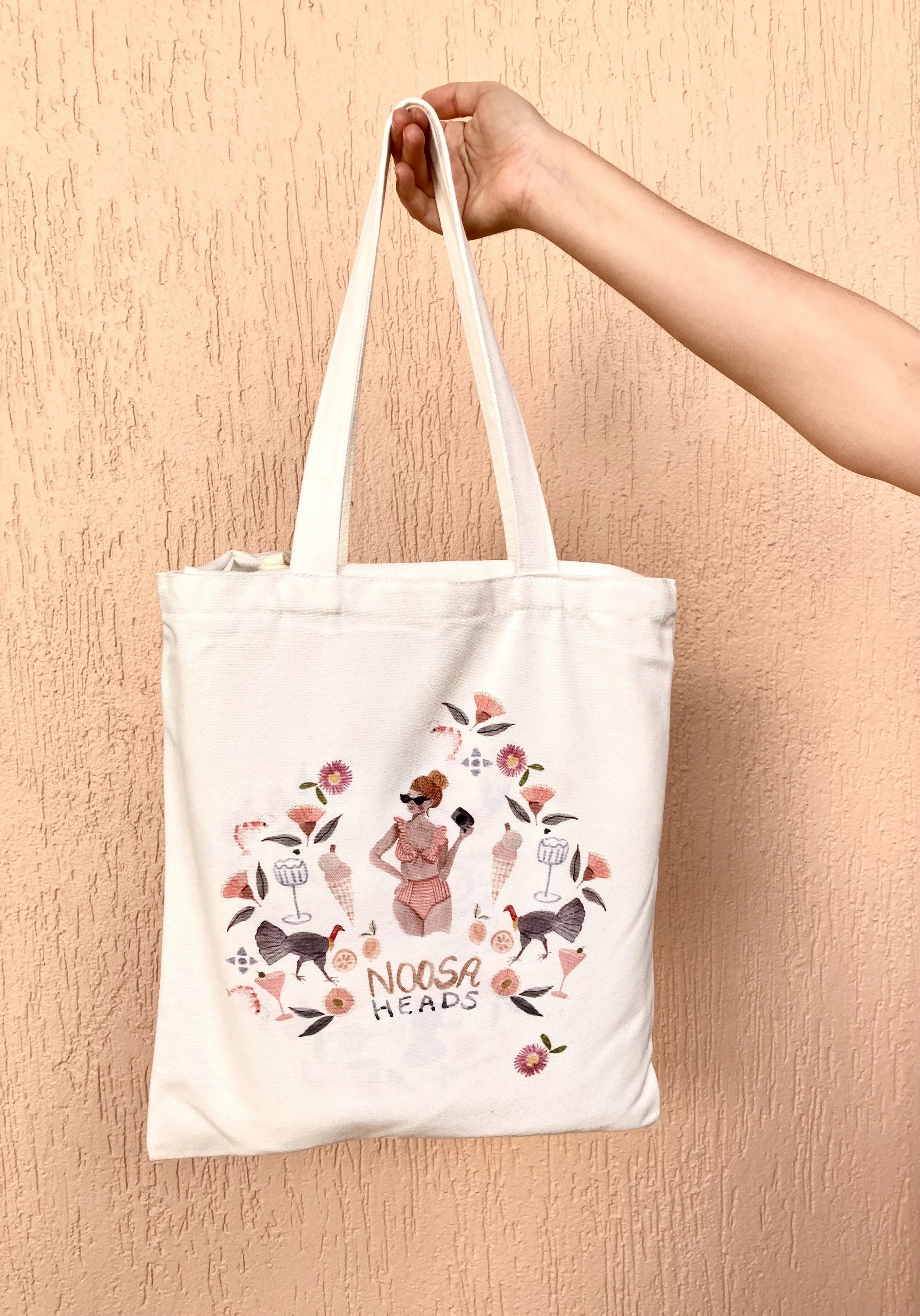 A white tote bag with a colorful illustration of a woman with sunglasses, surrounded by flowers, birds, ice cream cones, and a drink, with the text "NOSA HEADS" at the bottom, hanging from a hand against a peach-colored textured wall.
