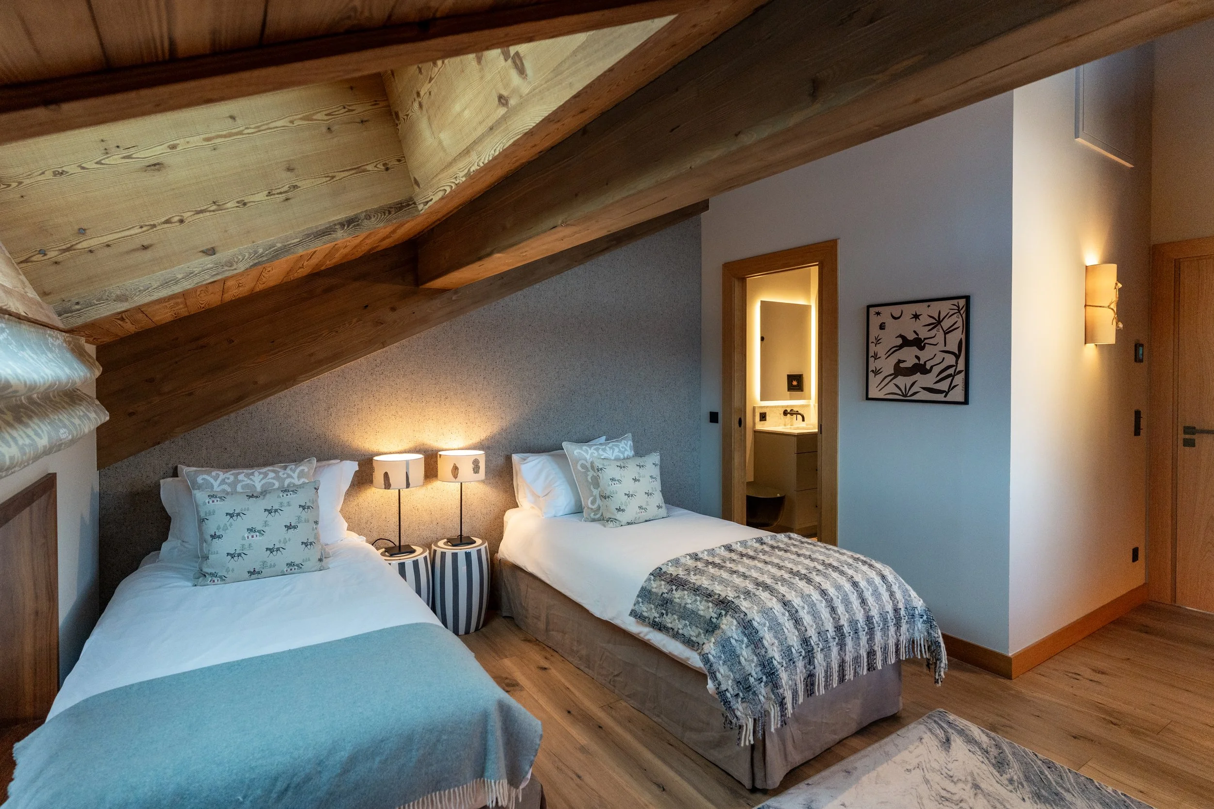 Cozy rustic bedroom with two twin beds, exposed wood beams, bedside lamps, and an adjoining bathroom