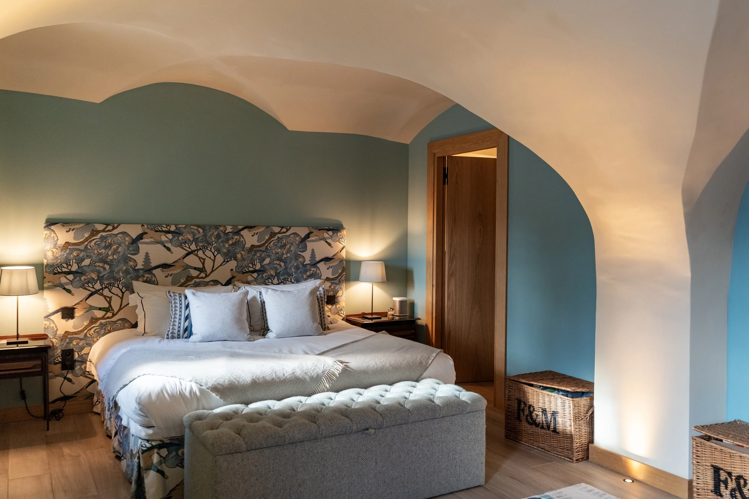 Cozy bedroom with floral upholstered headboard, white bed linens, blue walls, bedside lamps, wooden door, and wicker storage baskets.