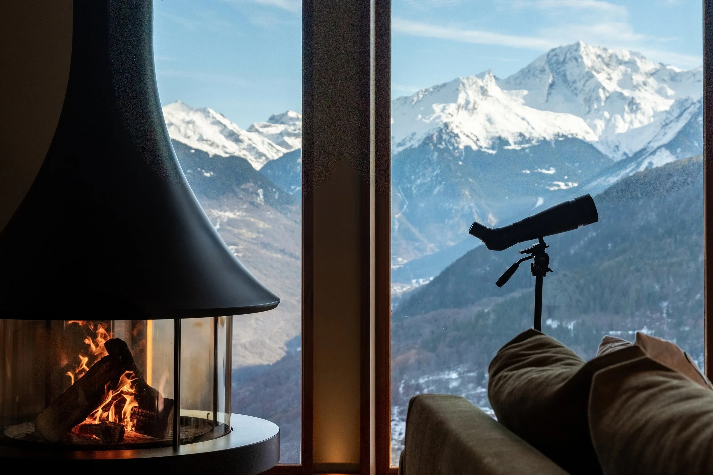 Cozy mountain-view room with a lit fireplace, telescope, and snow-covered Alps visible through floor-to-ceiling windows