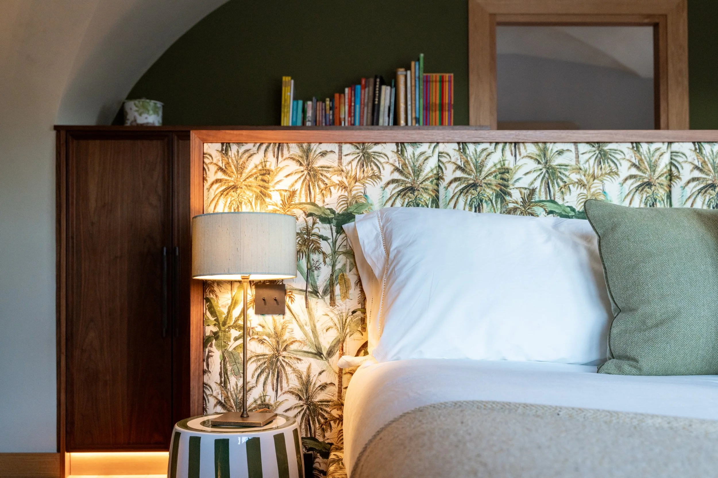 Cozy bedroom with palm tree wallpaper, white bedding, green pillow, bedside lamp, books, and dark wood cabinet