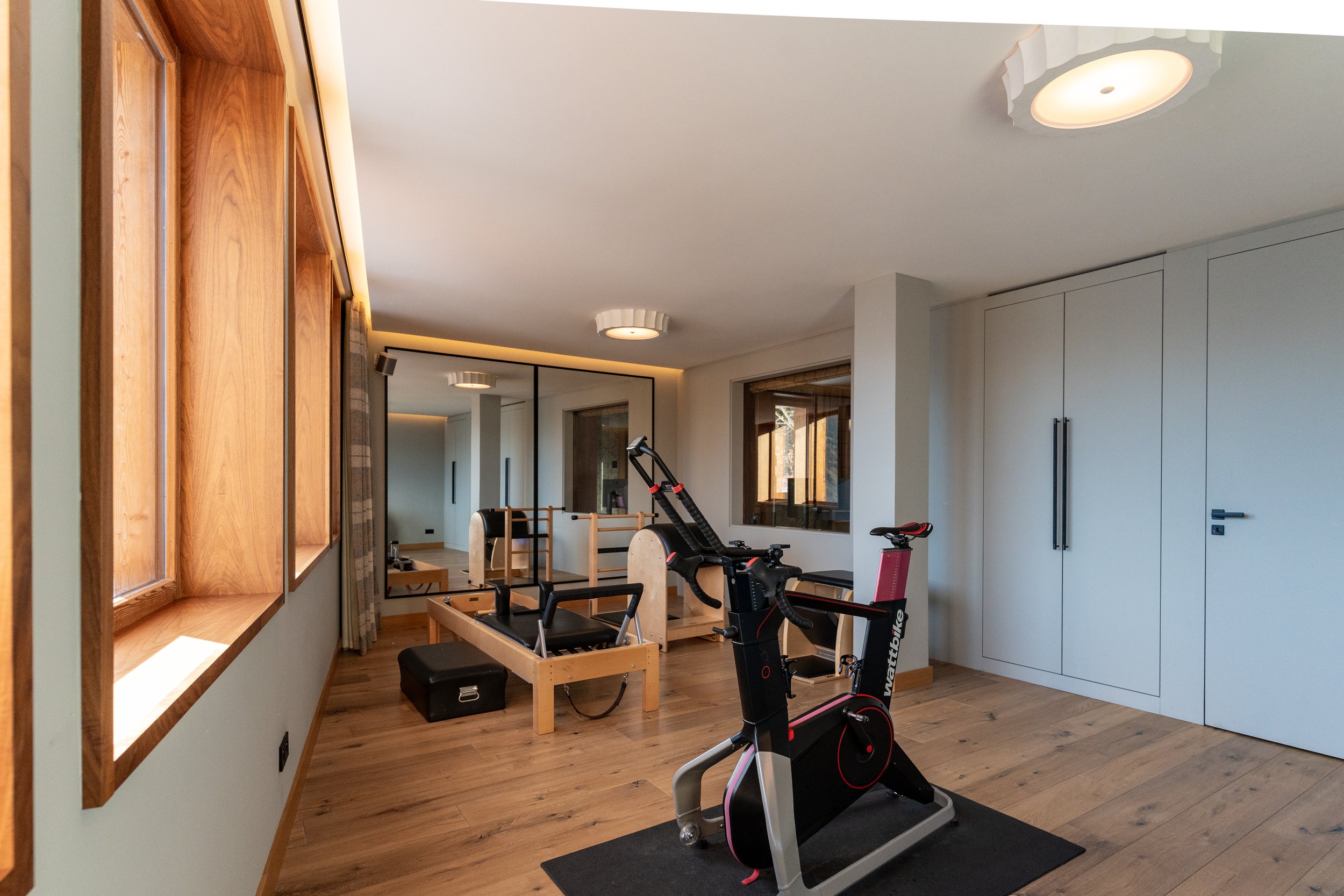 Modern home gym with stationary bike, Pilates reformer, mirrors, wood flooring, and built-in cabinets