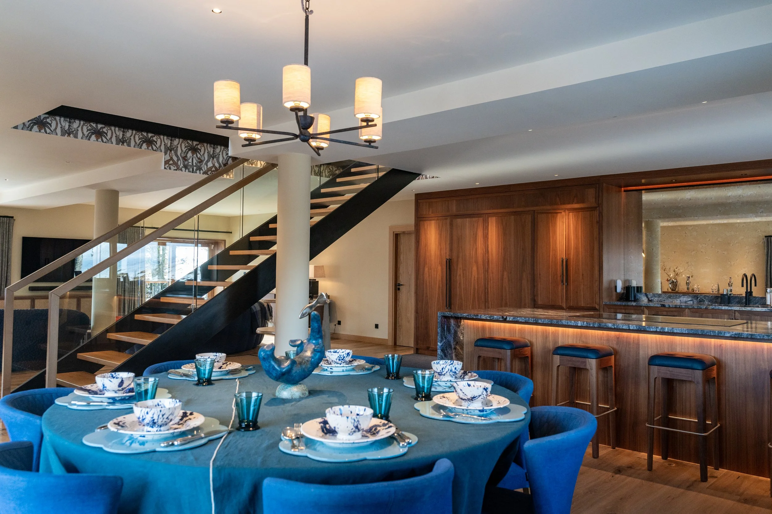 Modern open-plan dining room and kitchen with a blue-set dining table, contemporary staircase, wood cabinetry, and pendant chandelier