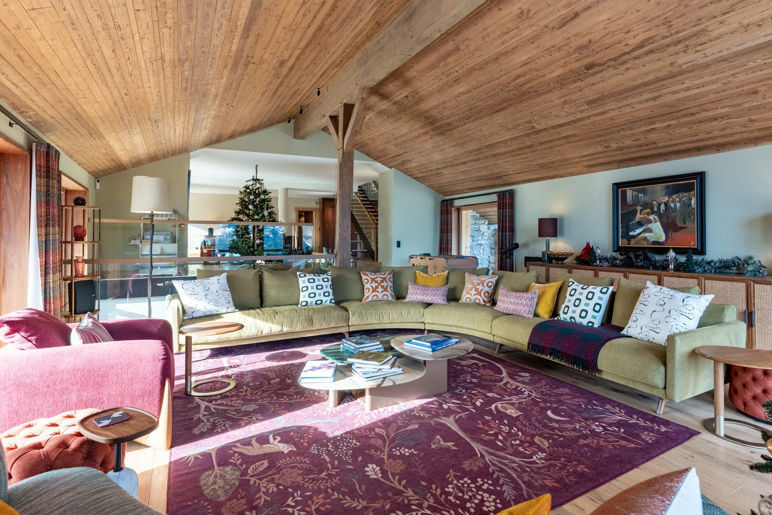 Spacious rustic living room with exposed wood ceilings, sectional sofa, patterned rug, coffee table, staircase, Christmas tree, and framed artwork