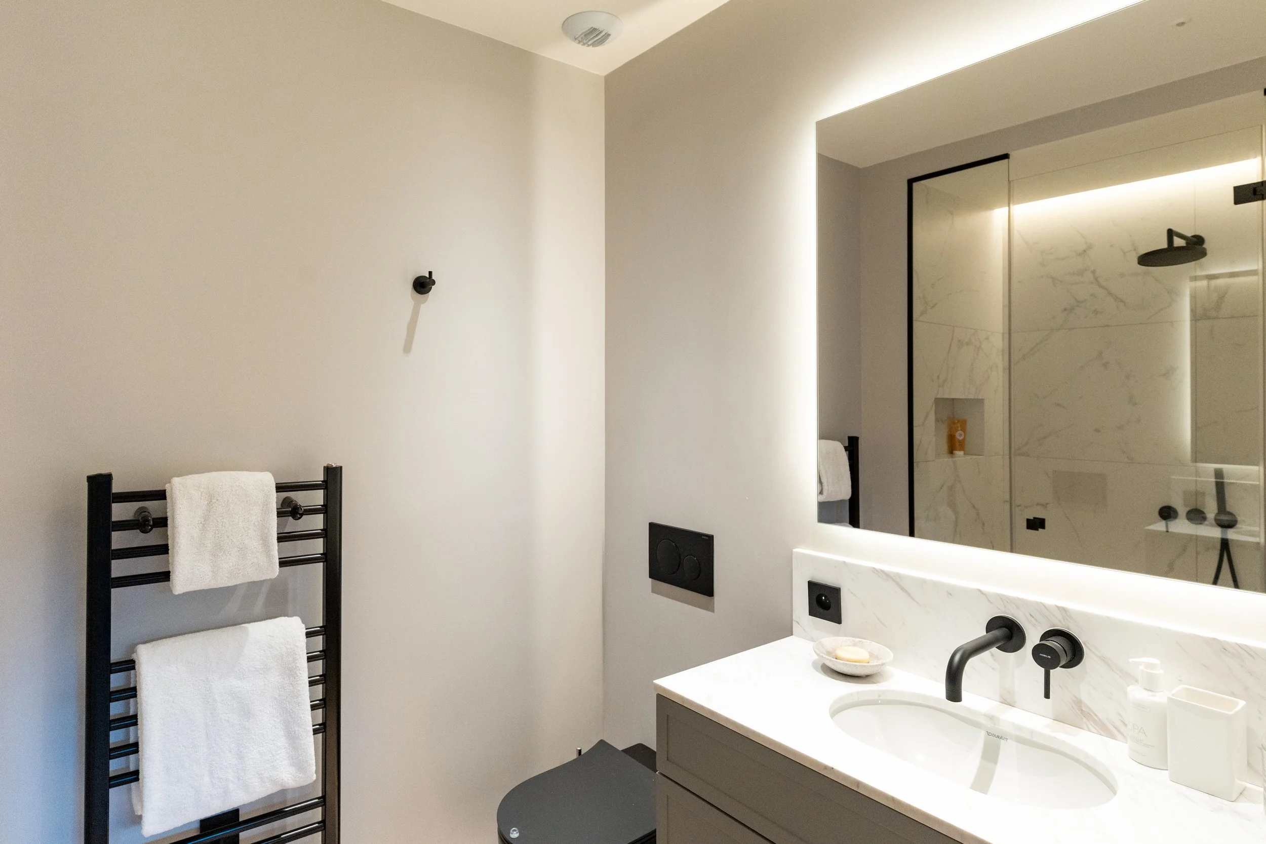 Modern bathroom with heated towel rack, marble vanity, illuminated mirror, and glass-enclosed walk-in shower