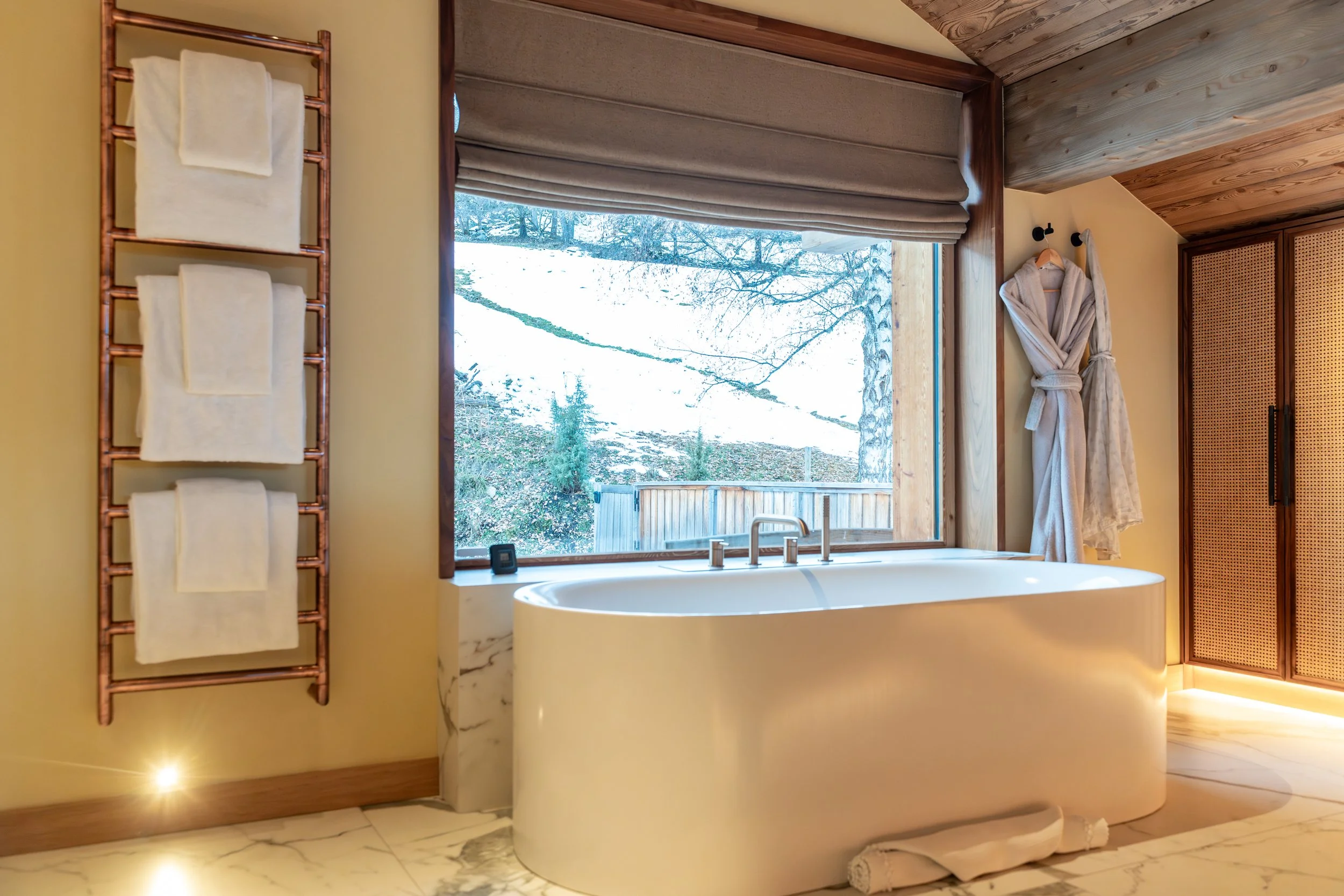 Luxury bathroom with freestanding bathtub, outdoor view, heated towel rack, robes, and wood cabinetry