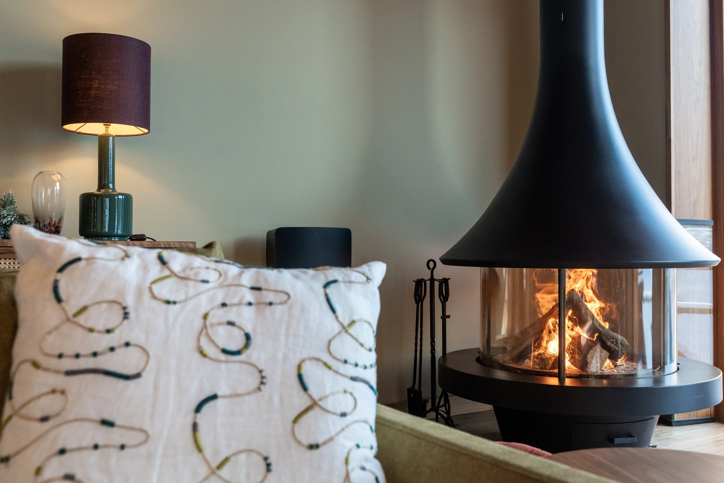 Cozy living room with a patterned pillow, table lamp, speaker, and a modern wood-burning fireplace.