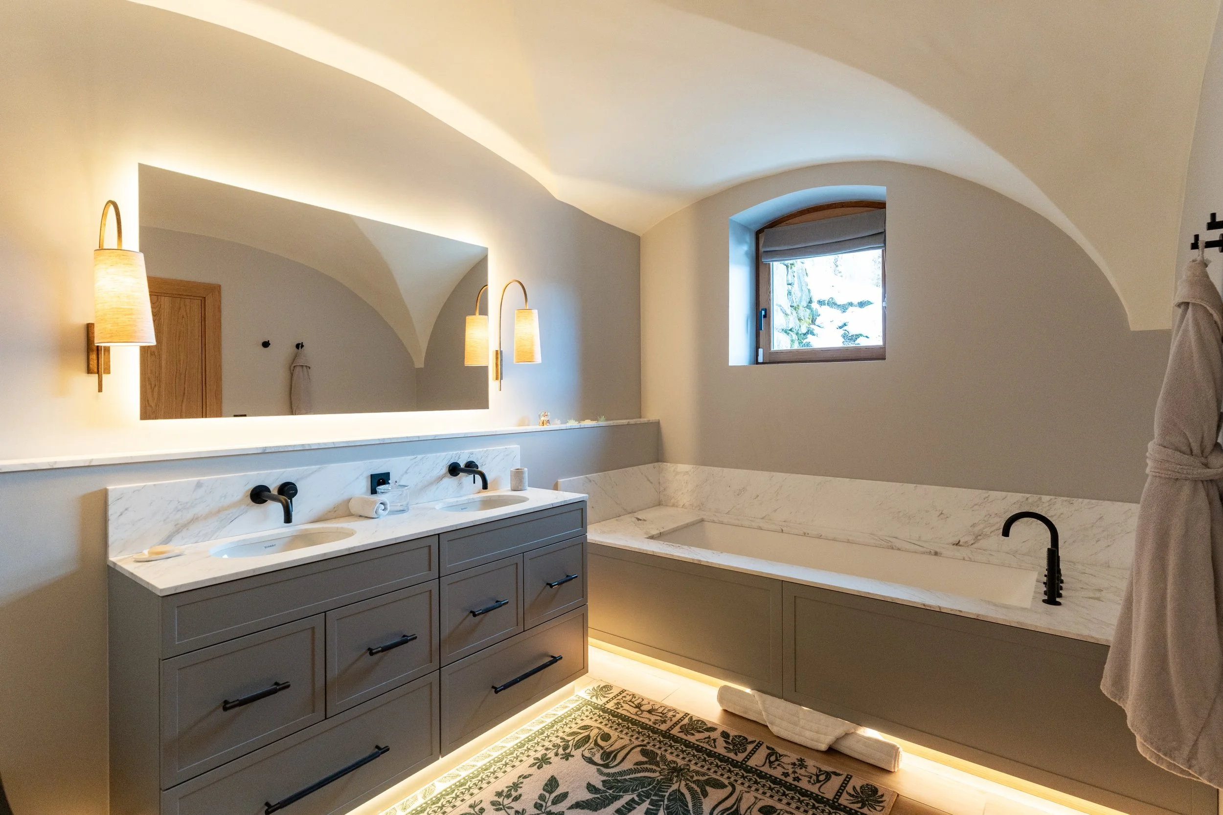 Elegant bathroom with gray double vanity, marble countertops, freestanding bathtub, arched window, and warm wall lighting