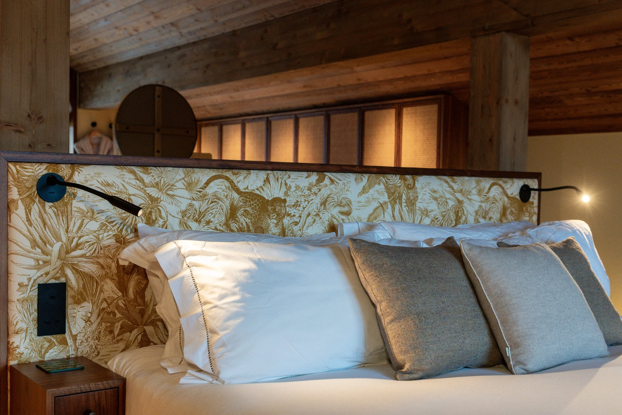 Cozy bedroom with a floral-patterned headboard, white bedding, gray pillows, wood-paneled walls, and reading lights
