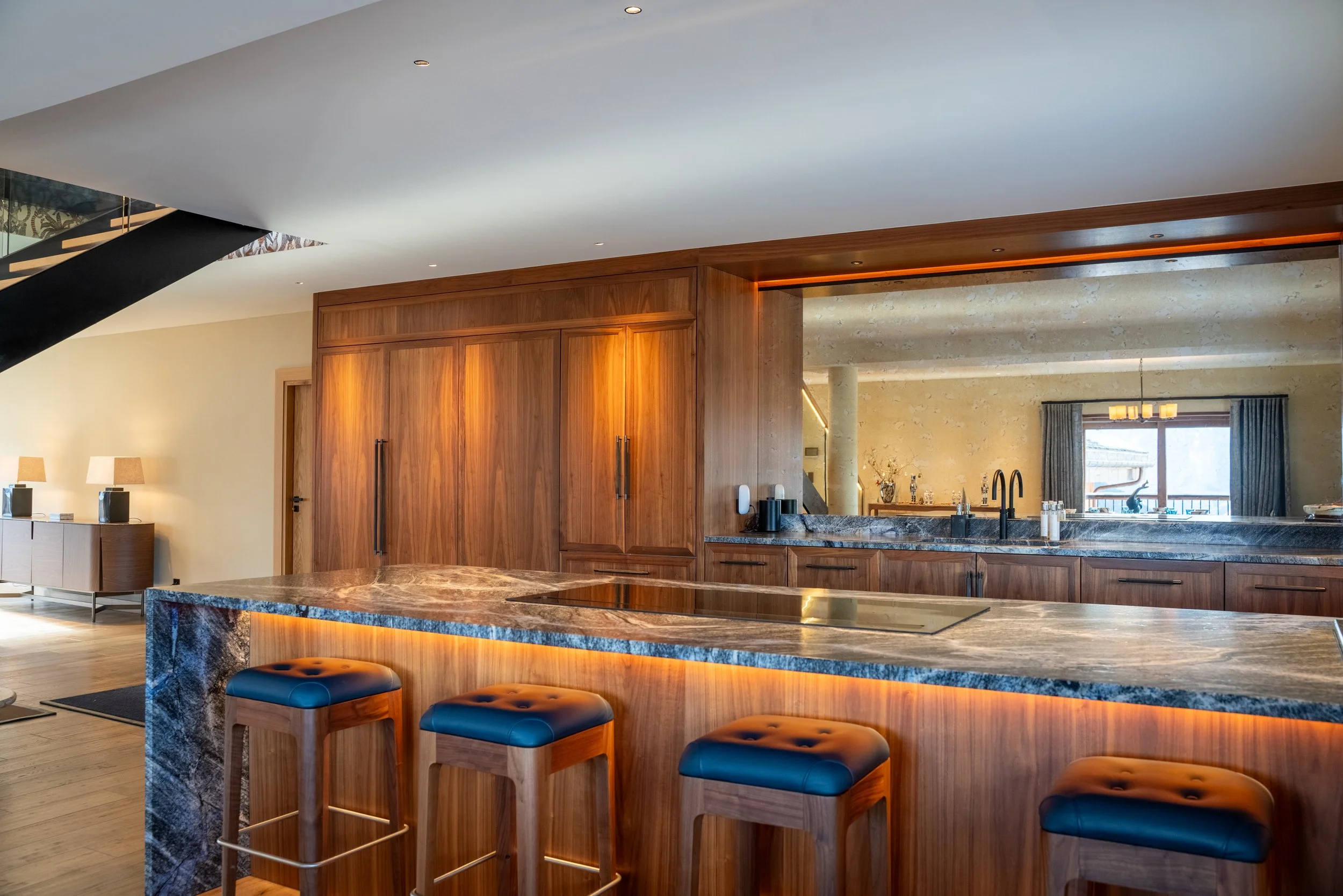 Luxury modern kitchen with walnut cabinetry, marble island, bar stools, and staircase