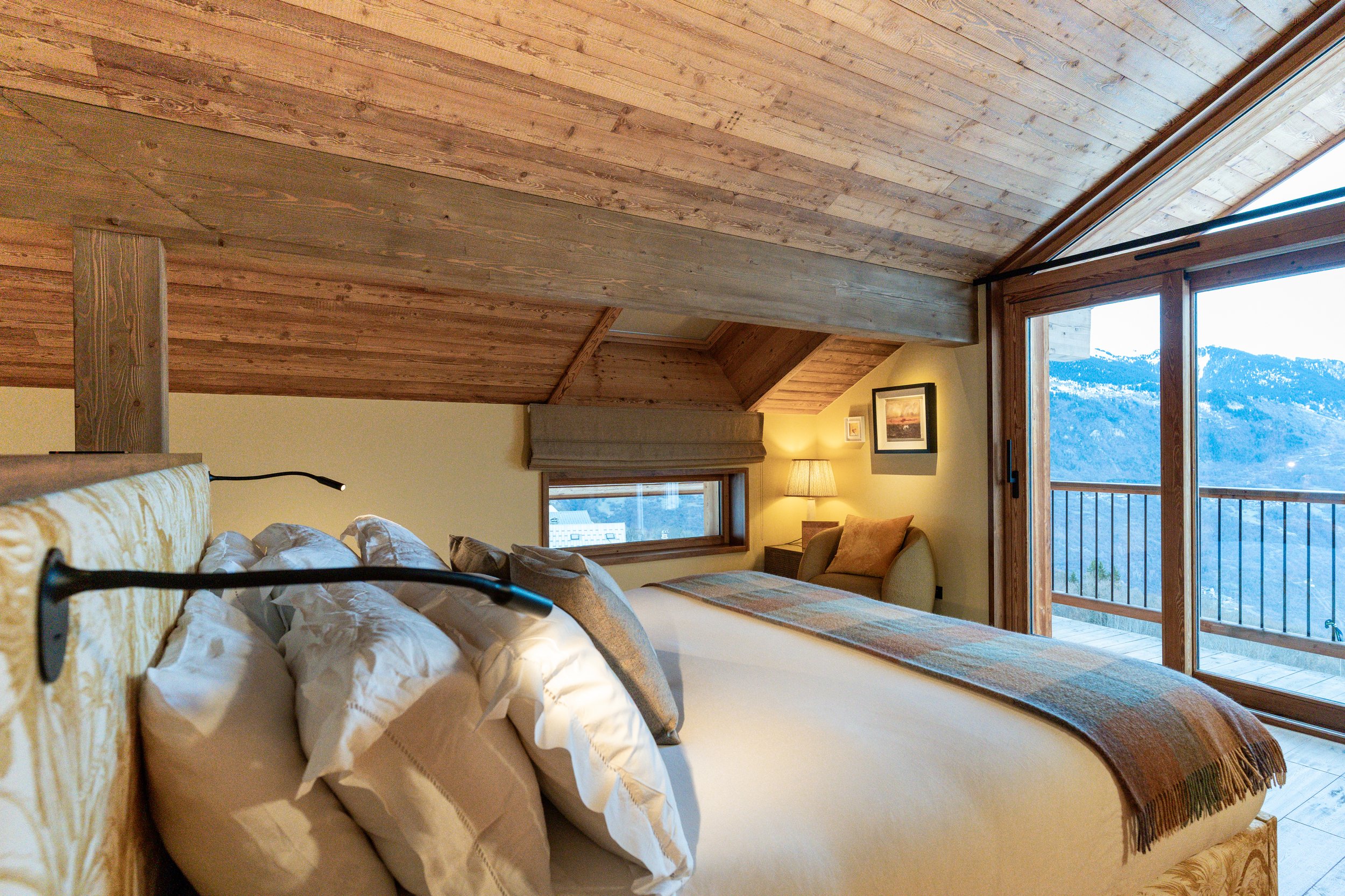 Cozy mountain living room with wood-paneled ceiling, sofas, armchair, lamp, and large windows overlooking a mountain landscape