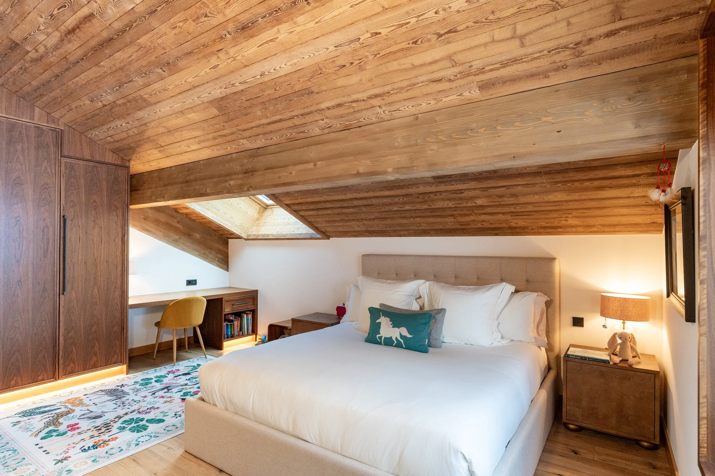 Cozy bedroom with a white upholstered bed, wood-paneled ceiling, built-in wardrobe, desk, skylight, bedside lamp, and patterned rug