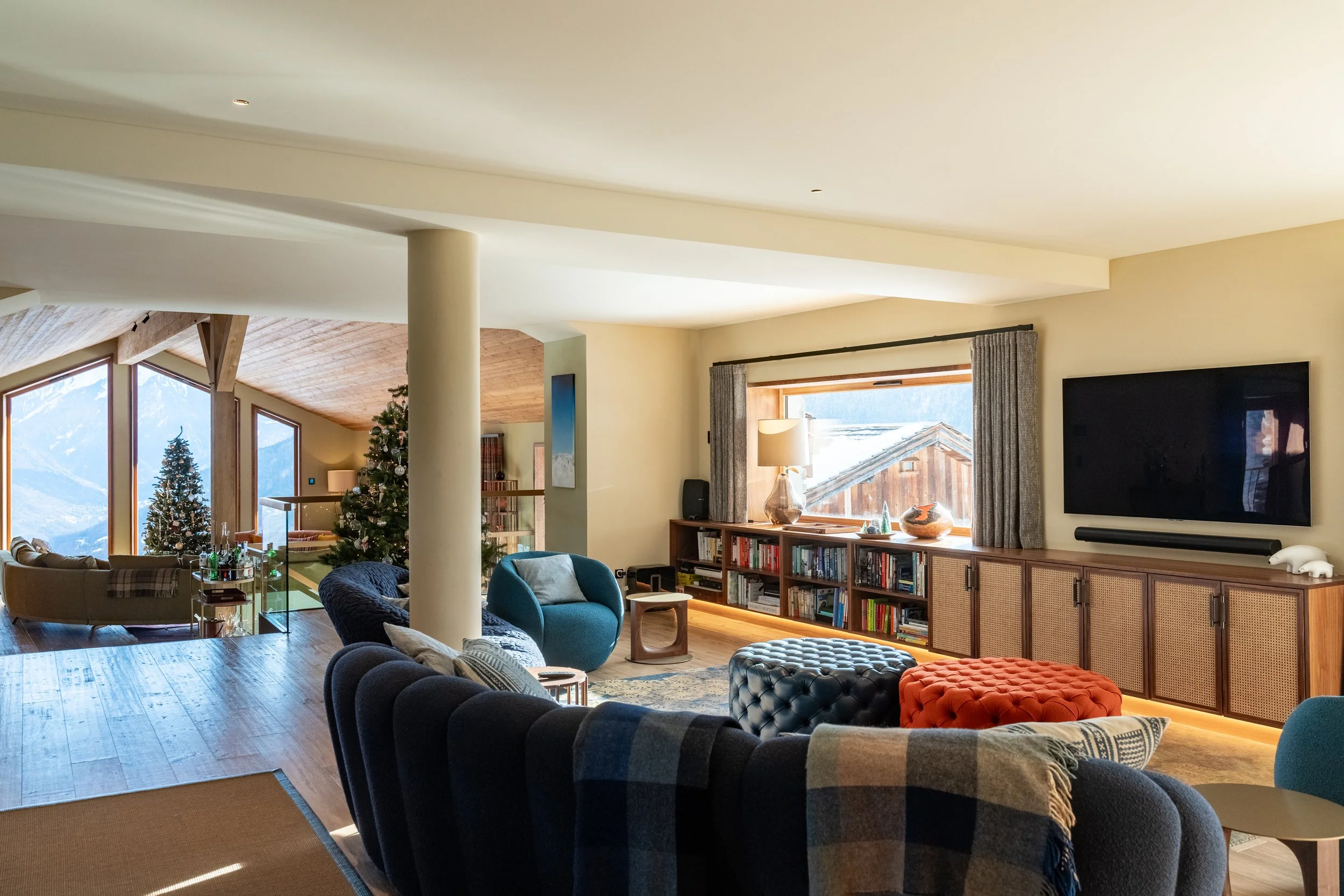 Mountain chalet living room with Christmas tree, large windows, blue sofas, colorful ottomans, bookshelves, and wall-mounted TV