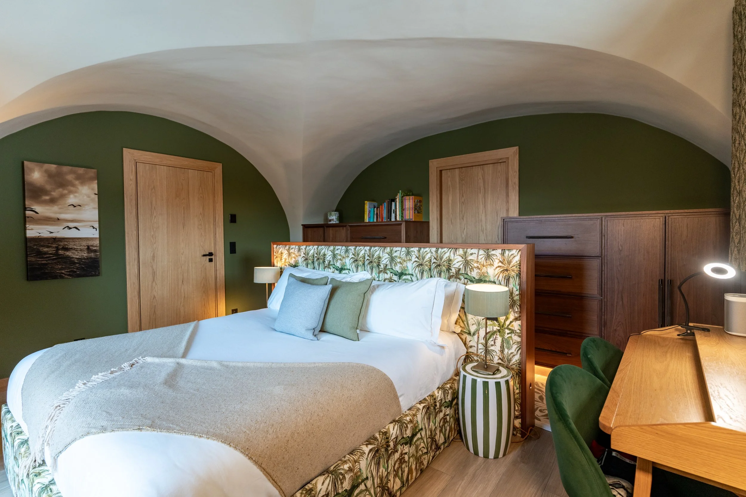 Stylish bedroom with a palm-print upholstered bed, green walls, wood furniture, desk, and arched ceiling