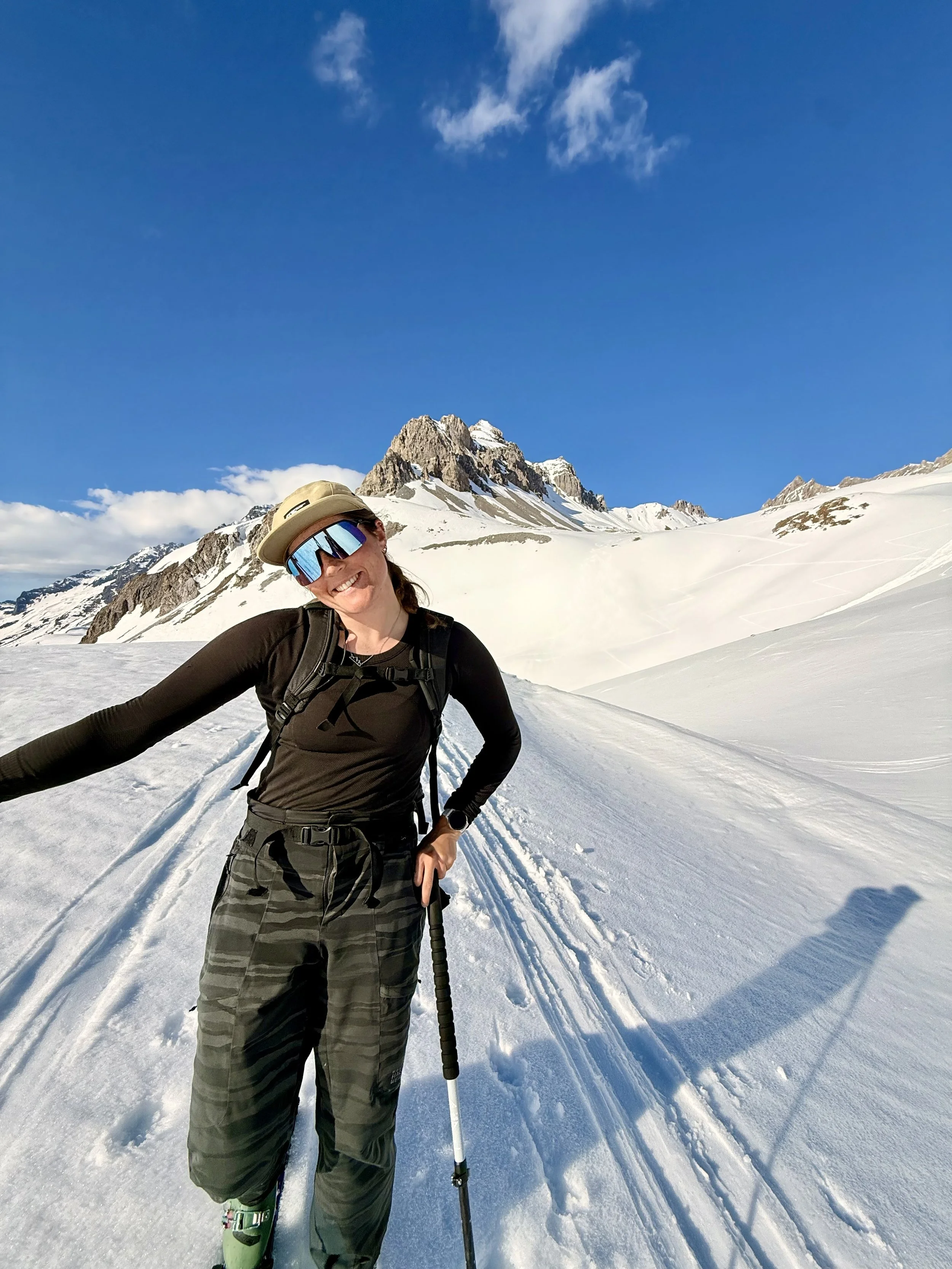 A smiling woman wearing sunglasses, a tan hat, a black long-sleeve top, green camouflage snow pants, and skis, standing in a snowy mountain landscape with a tall, rugged snow-covered mountain and a blue sky with some clouds in the background.