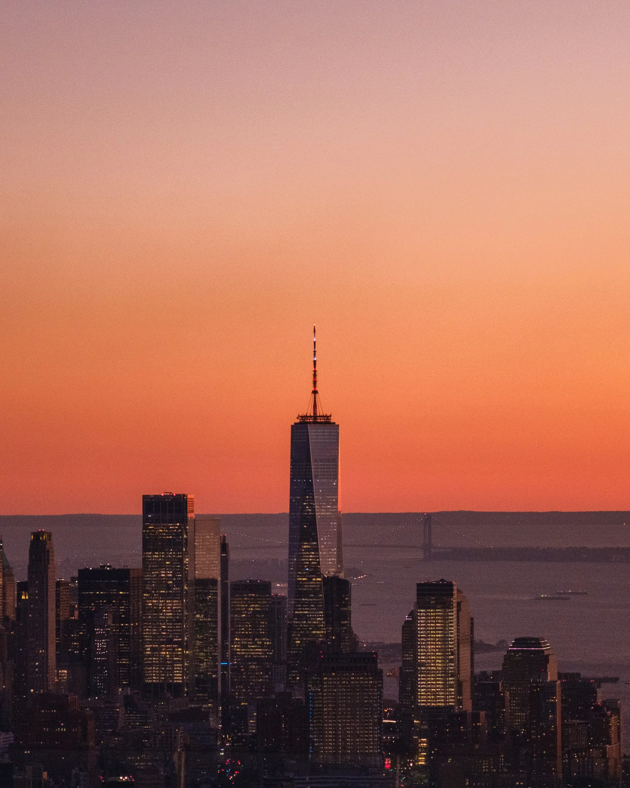 Skyline of a city at sunset with tall buildings, including one with a spire, and a bridge over water in the background.