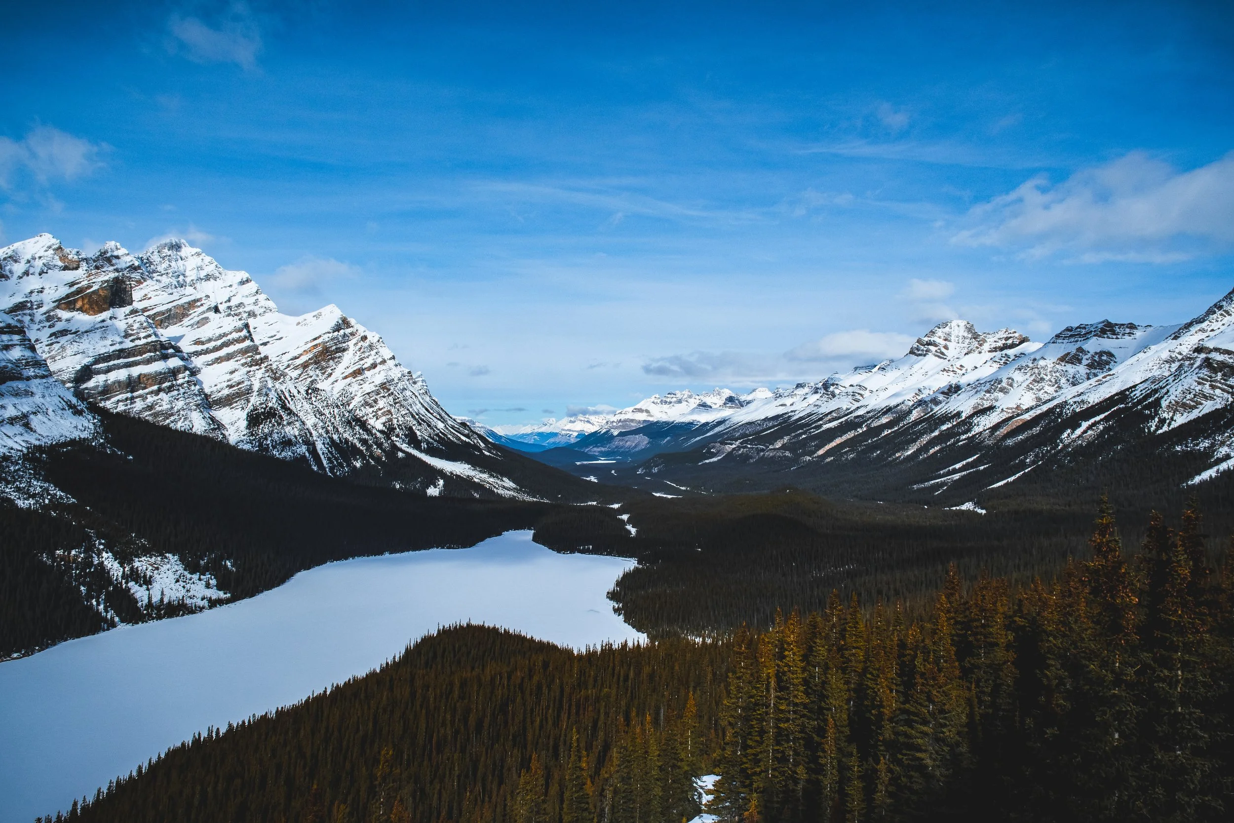 Snow-covered mountain peaks against a blue sky with some clouds, a lake at the base, and a dense forest of trees in the foreground.