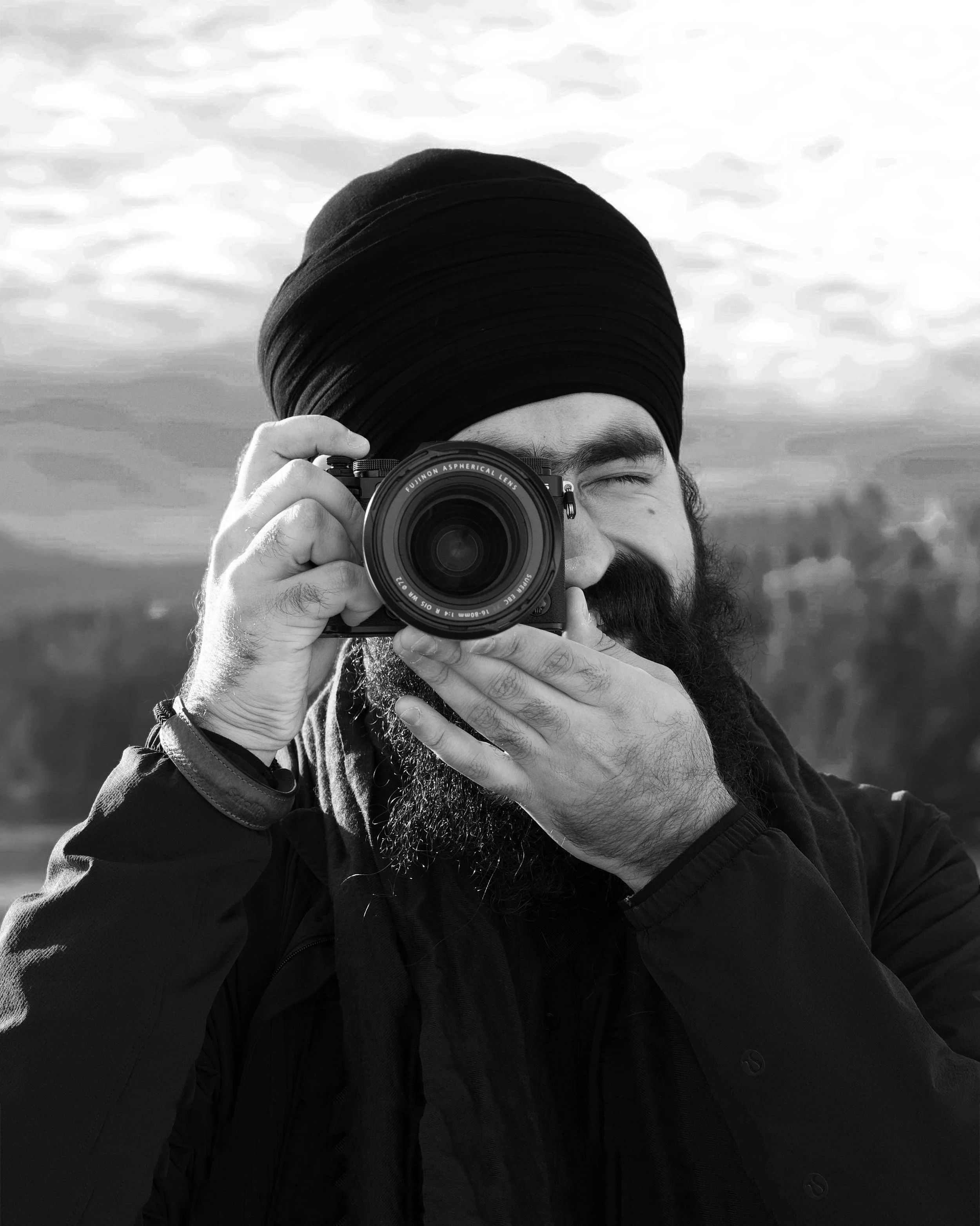 A man with a beard and glasses, wearing a black turban, taking a selfie outdoors with a camera, against a cloudy sky background.
