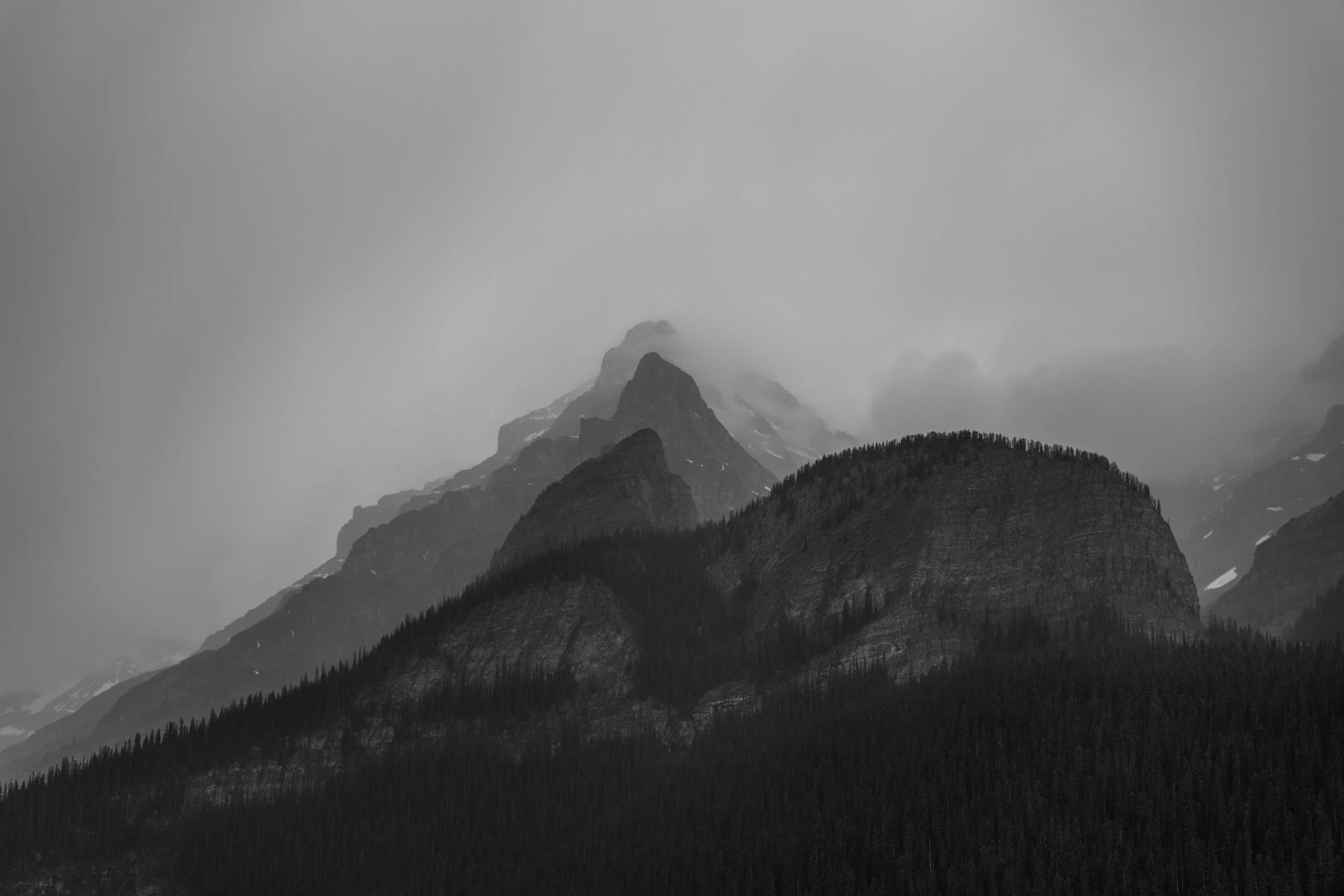 Black and white photograph of mountain peaks with cloudy sky and dense forest at the base.