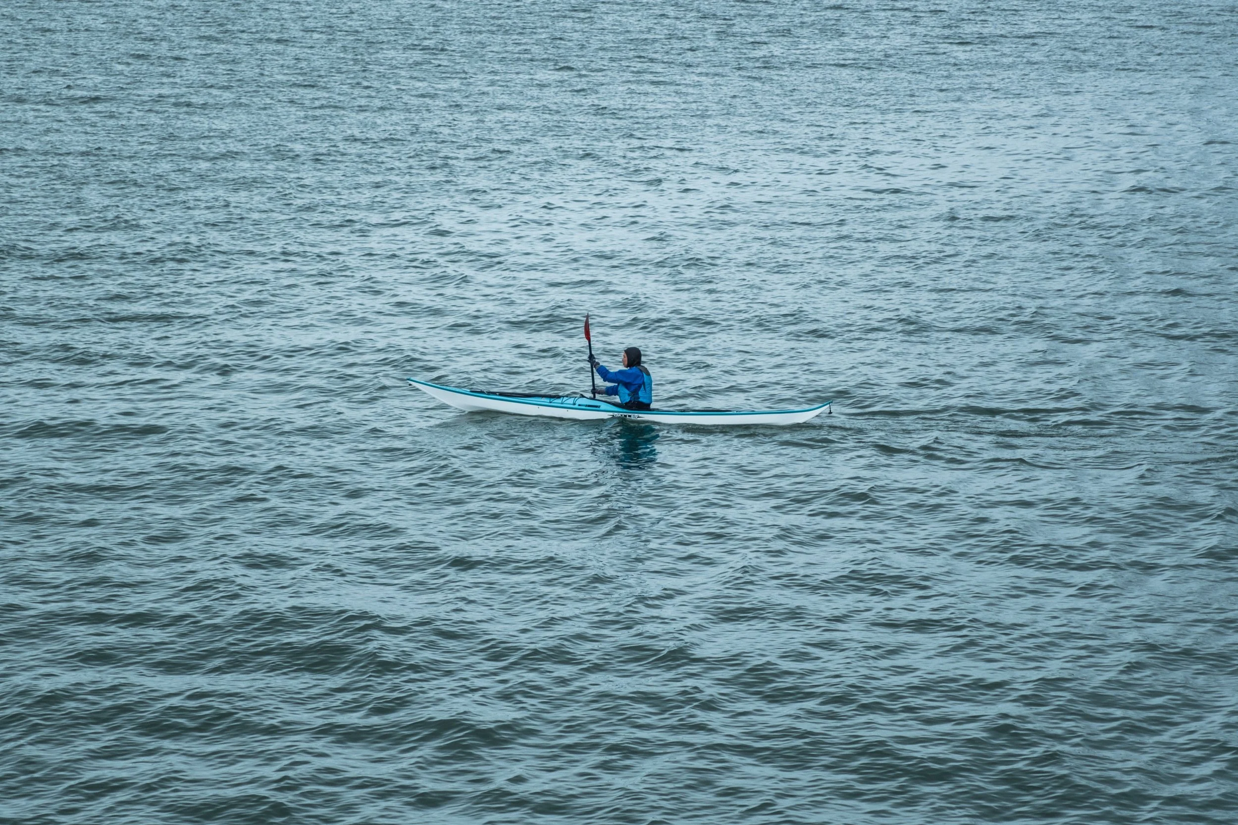 A person kayaking in a large body of water, wearing a blue jacket and a hood, holding a paddle and sitting in a small kayak.