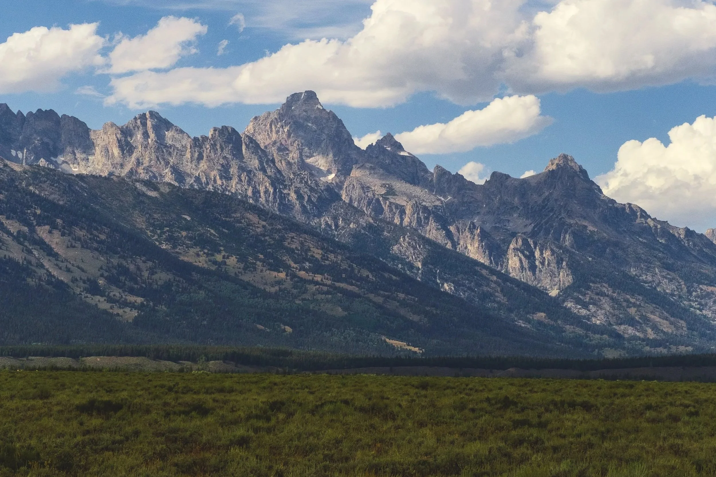 A mountain range with rugged, rocky peaks partly covered in snow, set against a partly cloudy blue sky. The foreground features a flat, grassy plain.