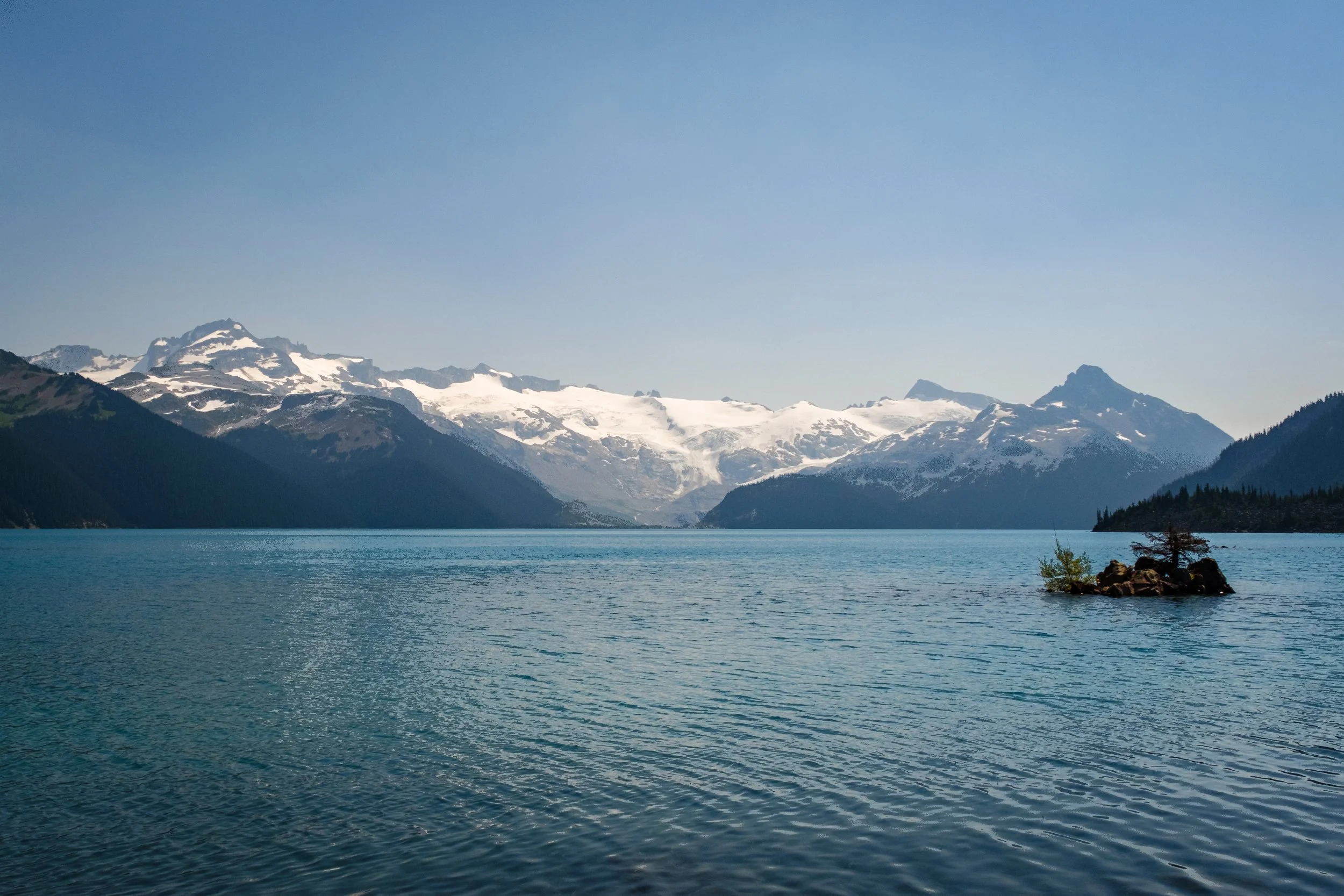 Mountain range with snow-capped peaks over a body of water, with a small rocky island with a tree in the foreground.