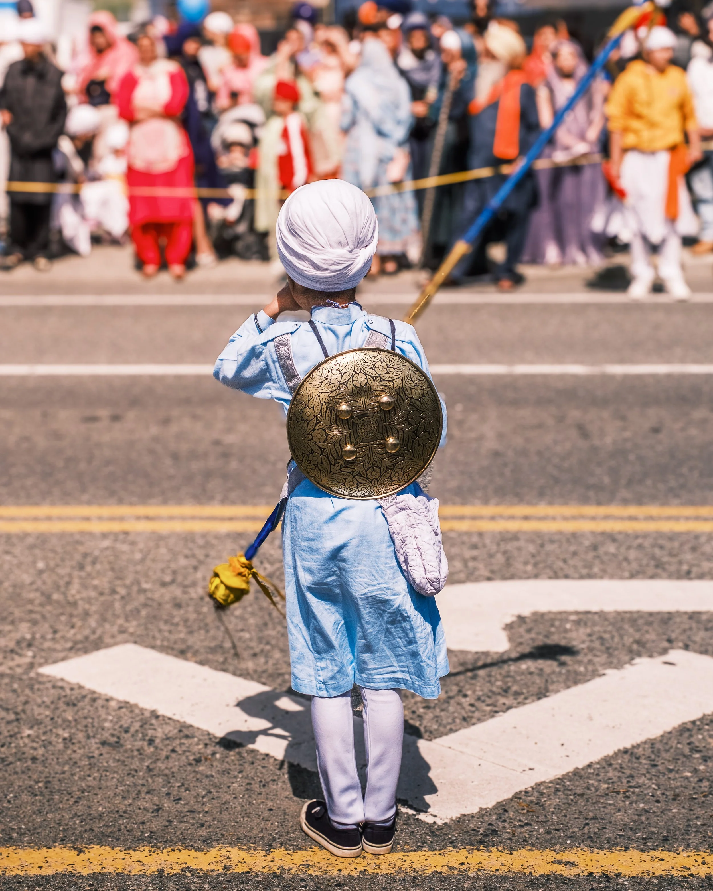 A young child dressed in a light blue costume with a white turban and holding a decorative shield stands on a crosswalk while people gather behind a yellow rope barrier, watching a parade or event.