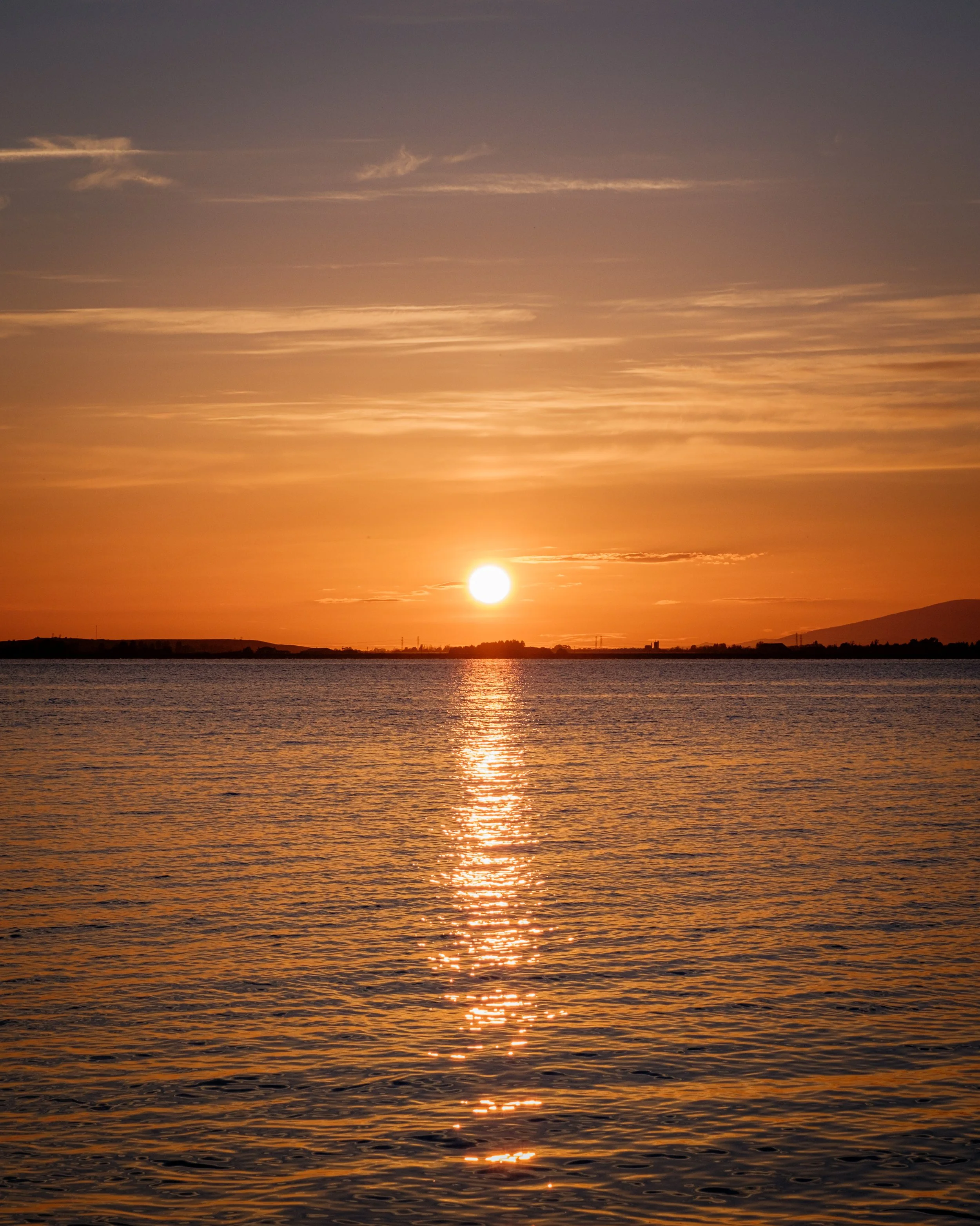 A sunset over a body of water with orange and yellow hues in the sky and the sun near the horizon, casting reflections on the water surface.