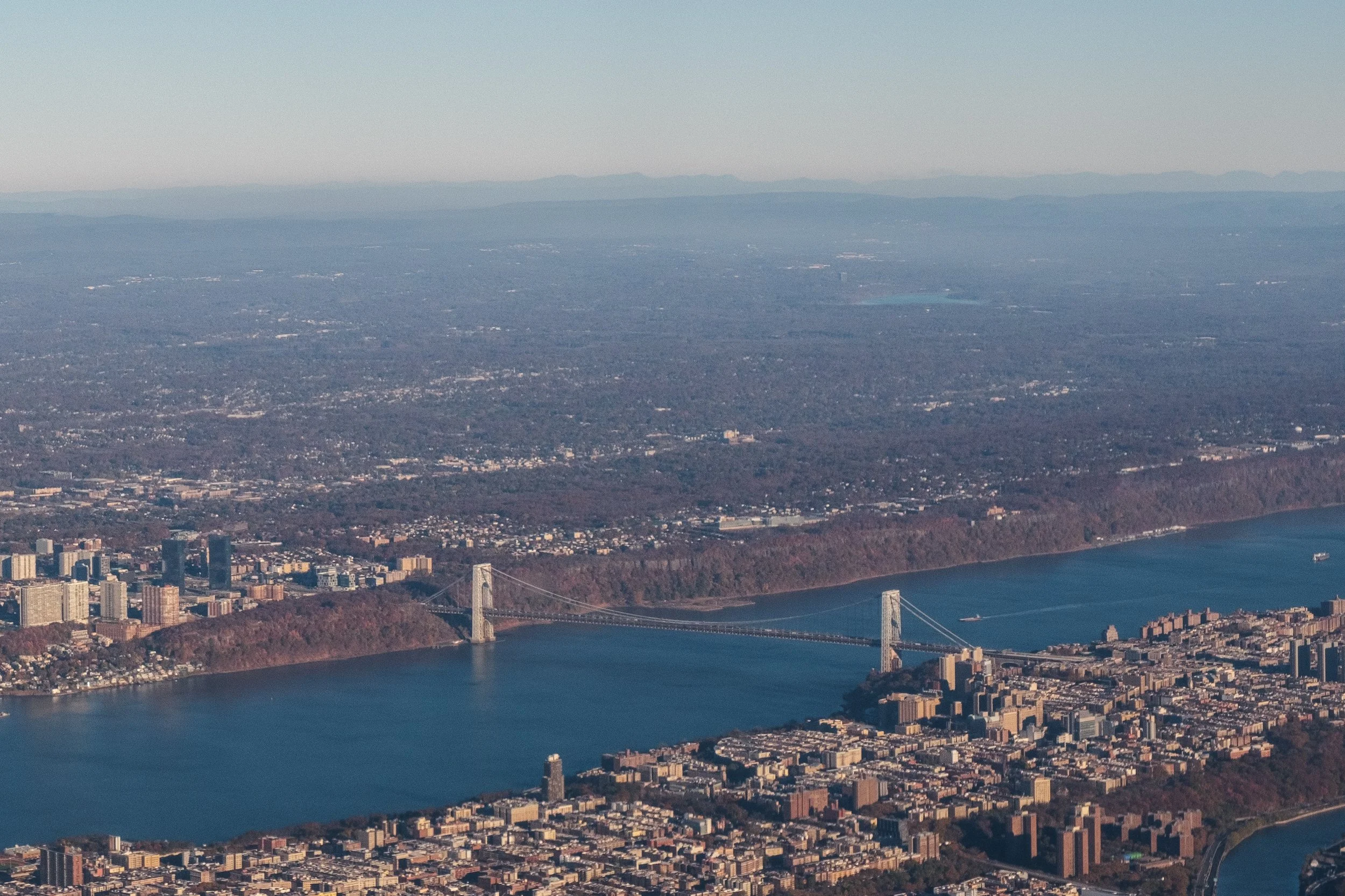 An aerial view of the San Francisco Bay Area, showing the Golden Gate Bridge, city buildings, water, and distant mountains.
