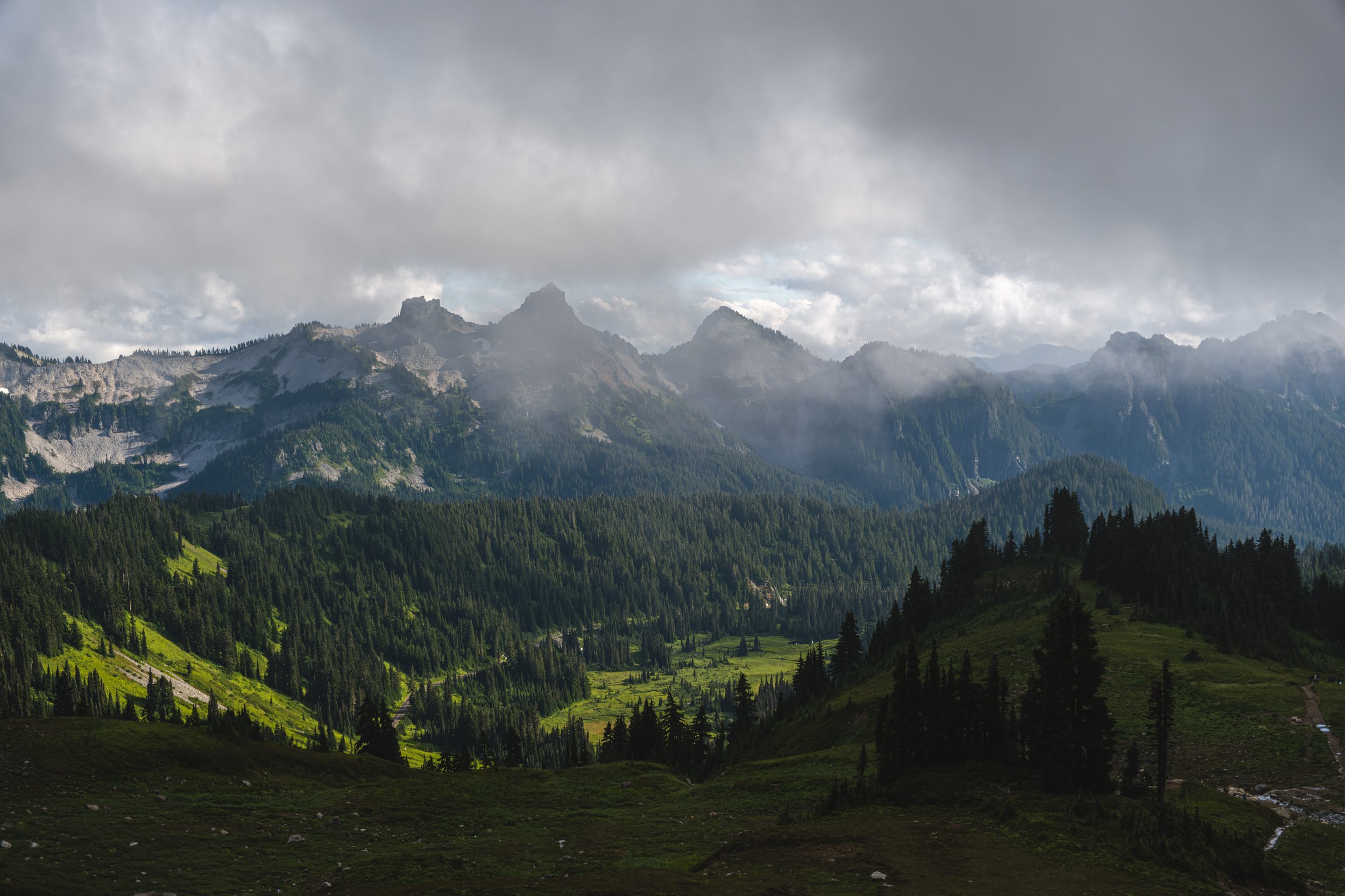 Mountain landscape with tall peaks, forested slopes, and cloudy sky.