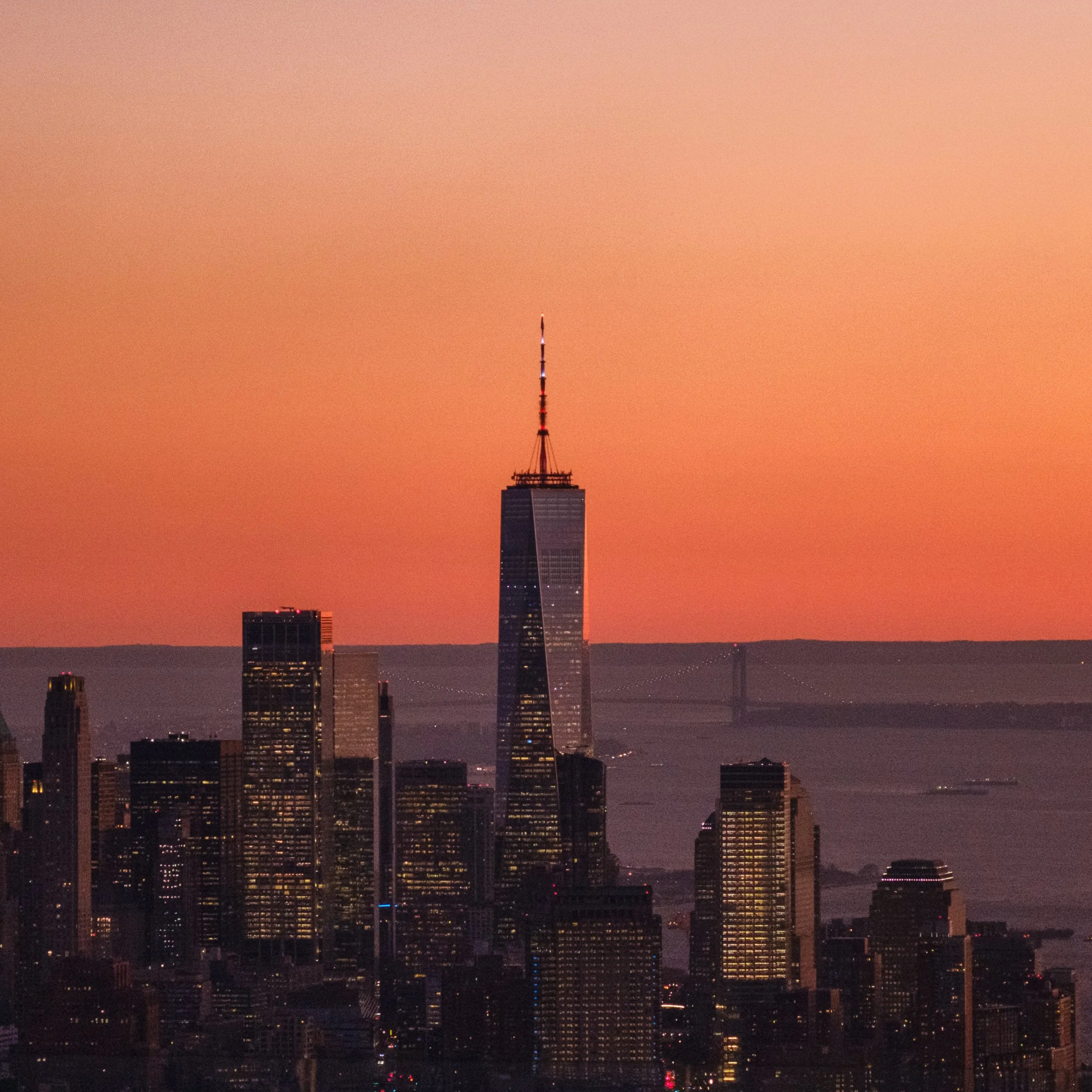 Silhouette of a city skyline with tall buildings, taken at dusk or dawn with an orange sky in the background.