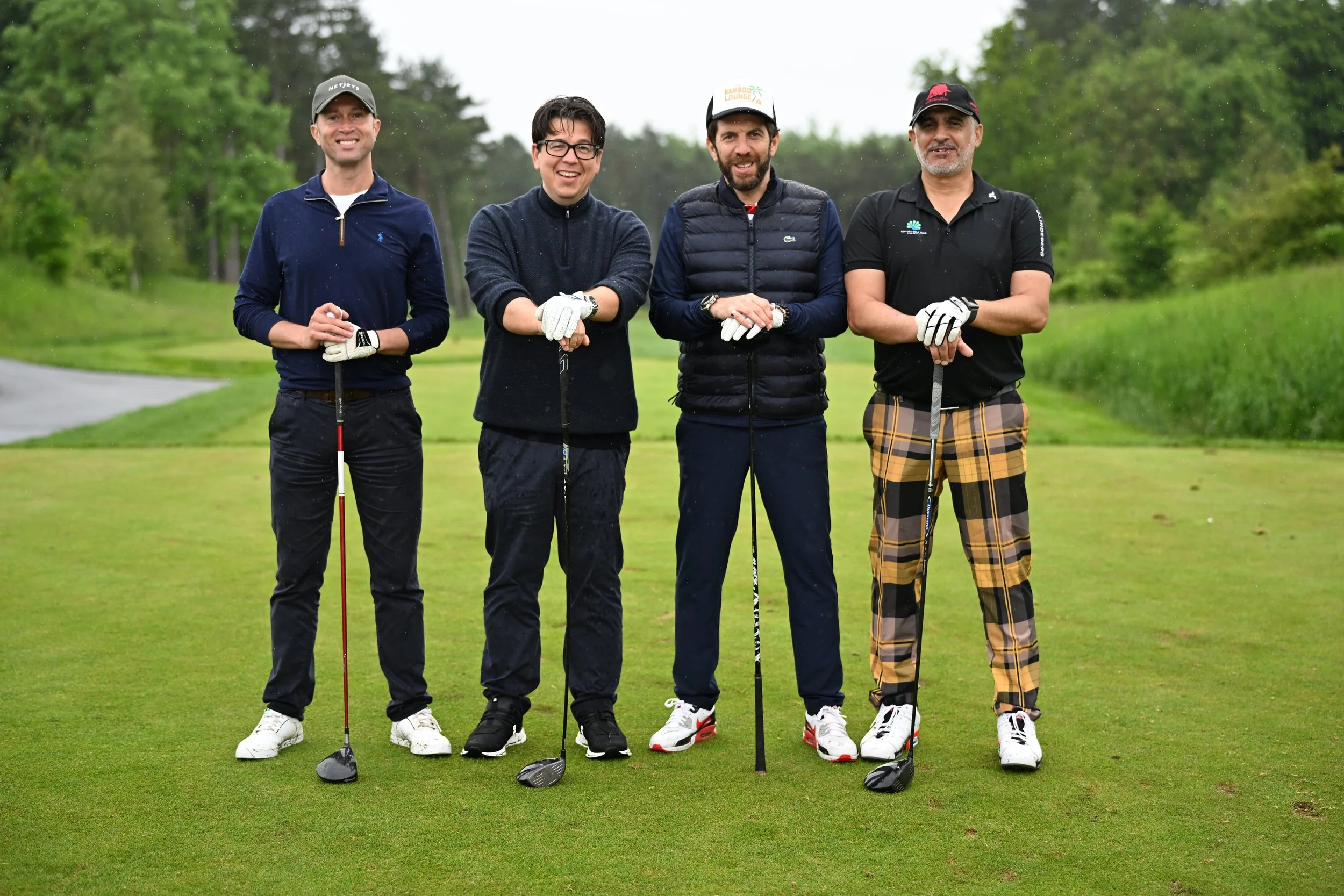 Four men standing on a golf course holding golf clubs, smiling, with trees and grass in the background, during a rainy day.