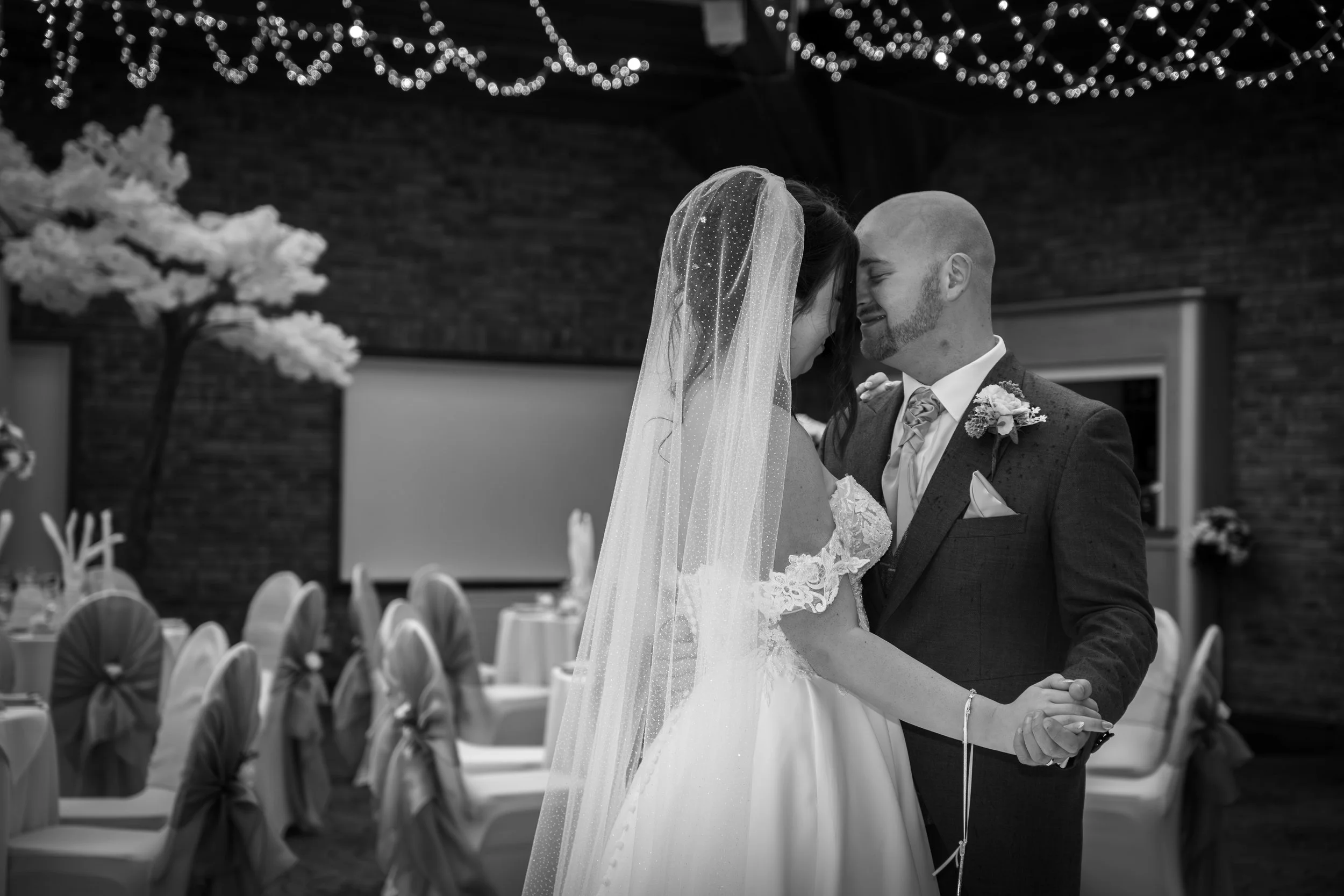 A black and white photo of a bride and groom dancing closely at their wedding reception. The bride wears a lace wedding dress and a veil, while the groom is in a suit with a boutonniere. The background shows decorated tables and string lights hanging from the ceiling.