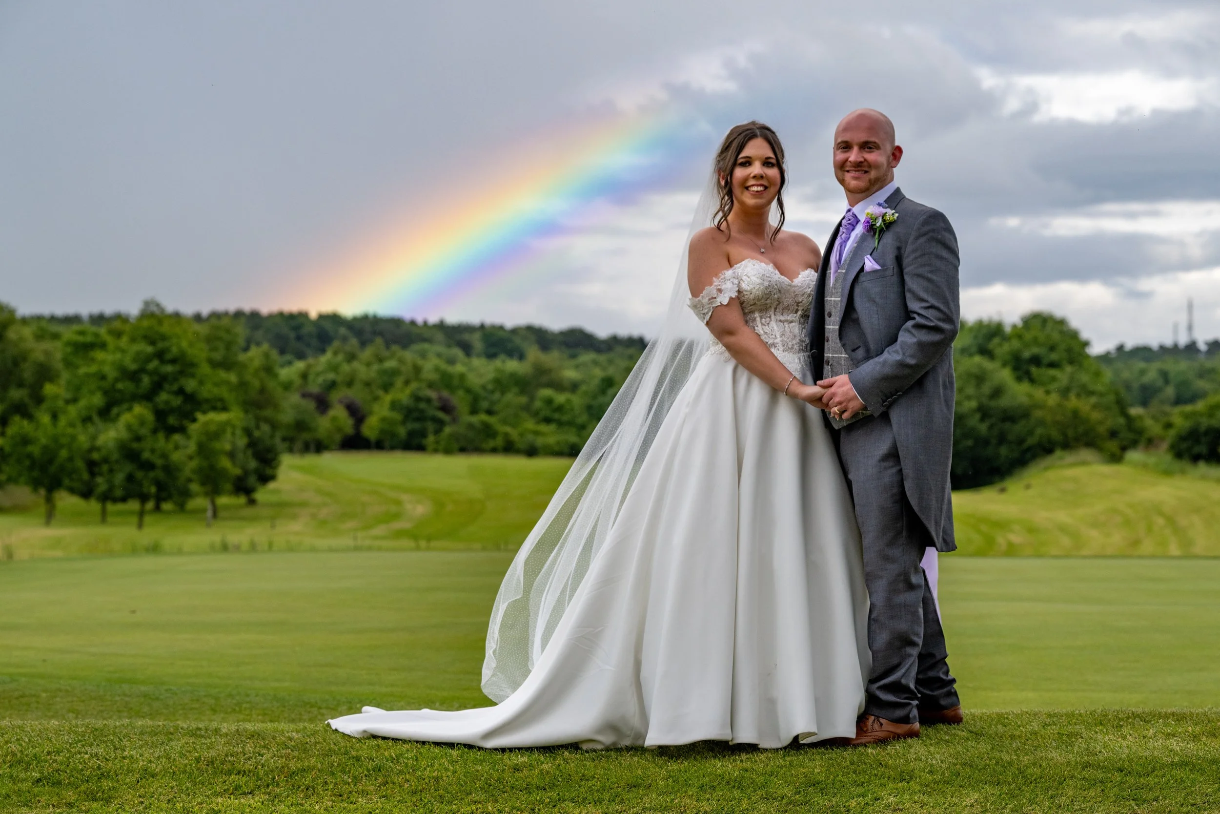 A bride and groom standing together outdoors on a grassy area with trees in the background. The bride is wearing a white lace wedding dress and veil, and the groom is dressed in a gray suit with a purple tie and boutonniere. There is a rainbow in the cloudy sky behind them.