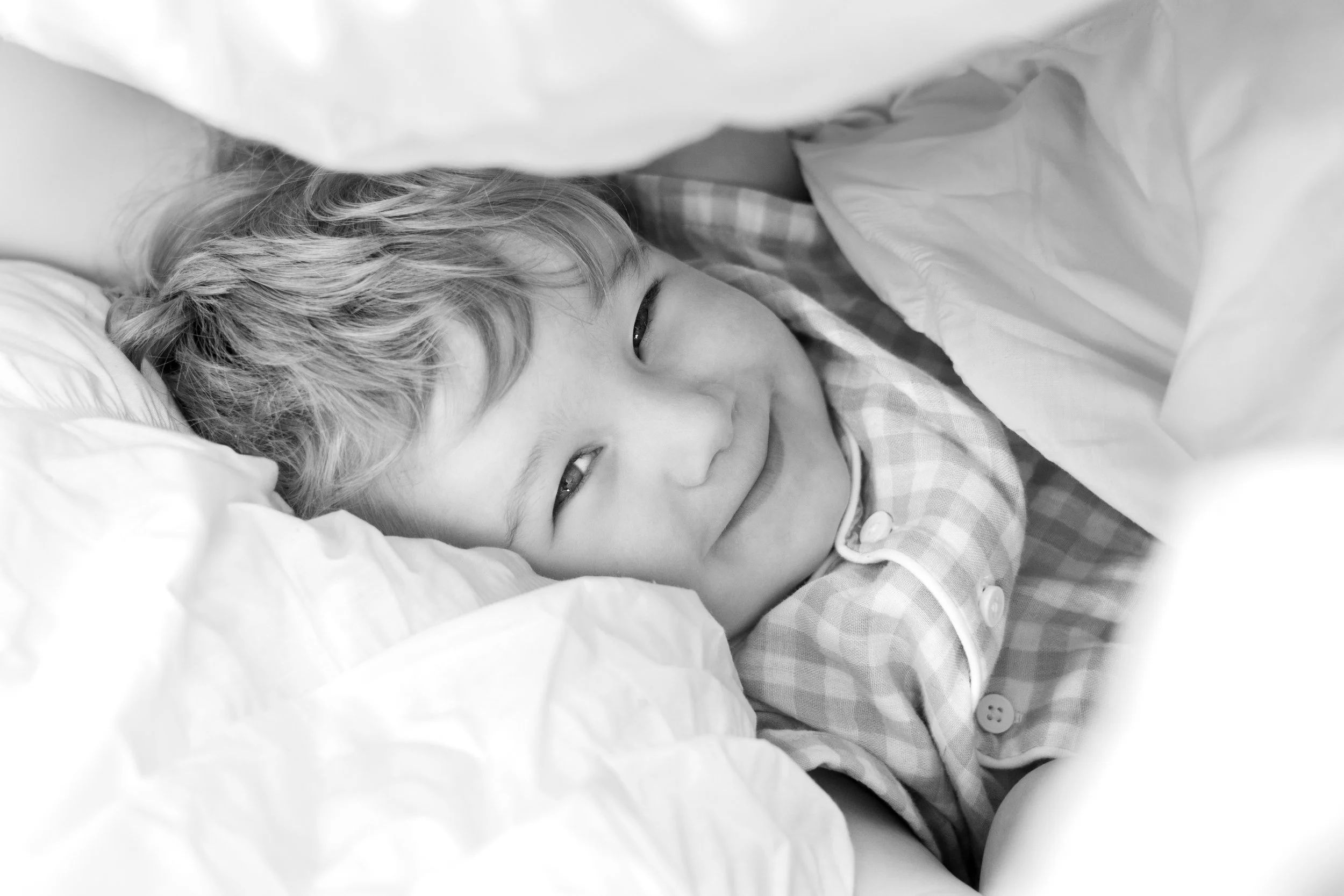 A young child with light-colored, wavy hair, lying in bed under a blanket, smiling with eyes partially closed.