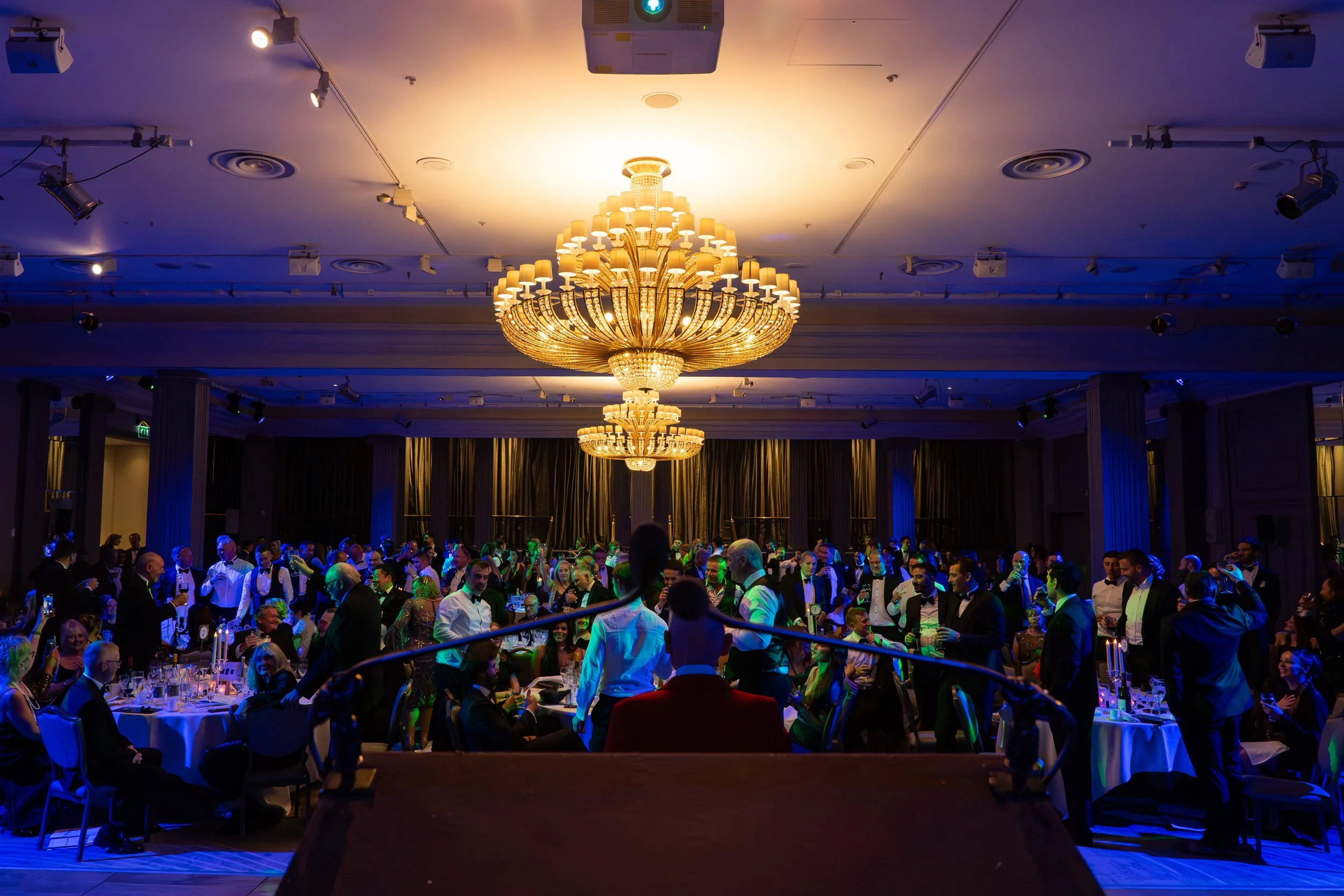 Elegant ballroom decorated for a formal event with a large ornate chandelier hanging from the ceiling, filled with many guests dressed in tuxedos and evening gowns, socializing at round tables with elegant table settings and candles.