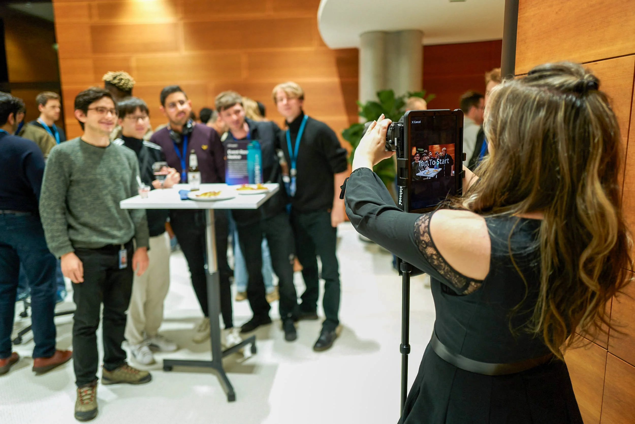 A woman with long hair taking a photo of a group of young men at a social event in a room with wooden walls and a potted plant.