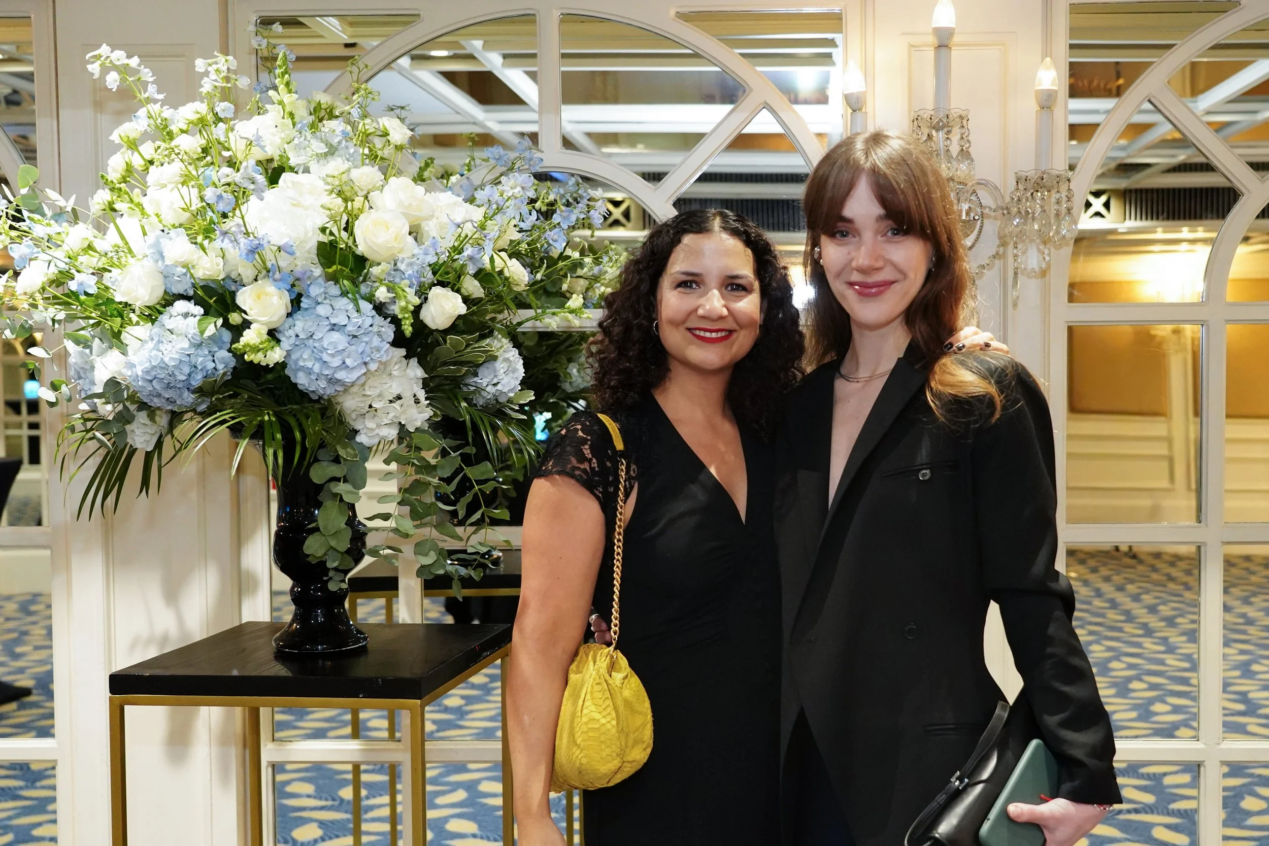 Two women smiling and standing next to a large floral arrangement of white and blue flowers in a black vase, in an elegant indoor setting with decorative lighting and mirrors.